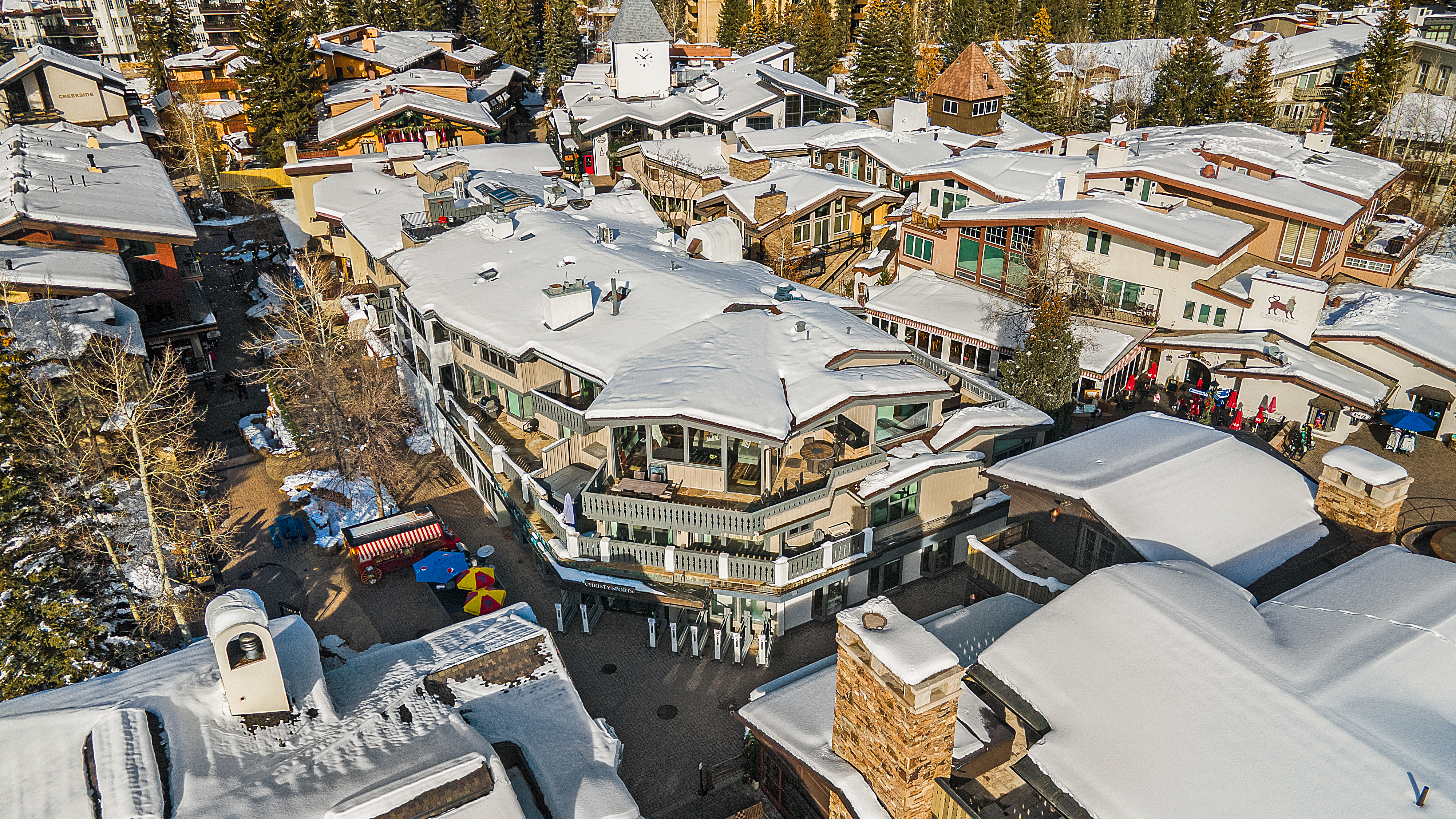 This aerial shot showcases a luxurious multi-story building with snow-covered roofs, surrounded by other buildings and trees, also covered in snow. The building features multiple balconies and a unique architectural design, suggesting a high-end residential or commercial property in a winter resort town. The perspective gives a comprehensive view of the property's setting and scale within the community.