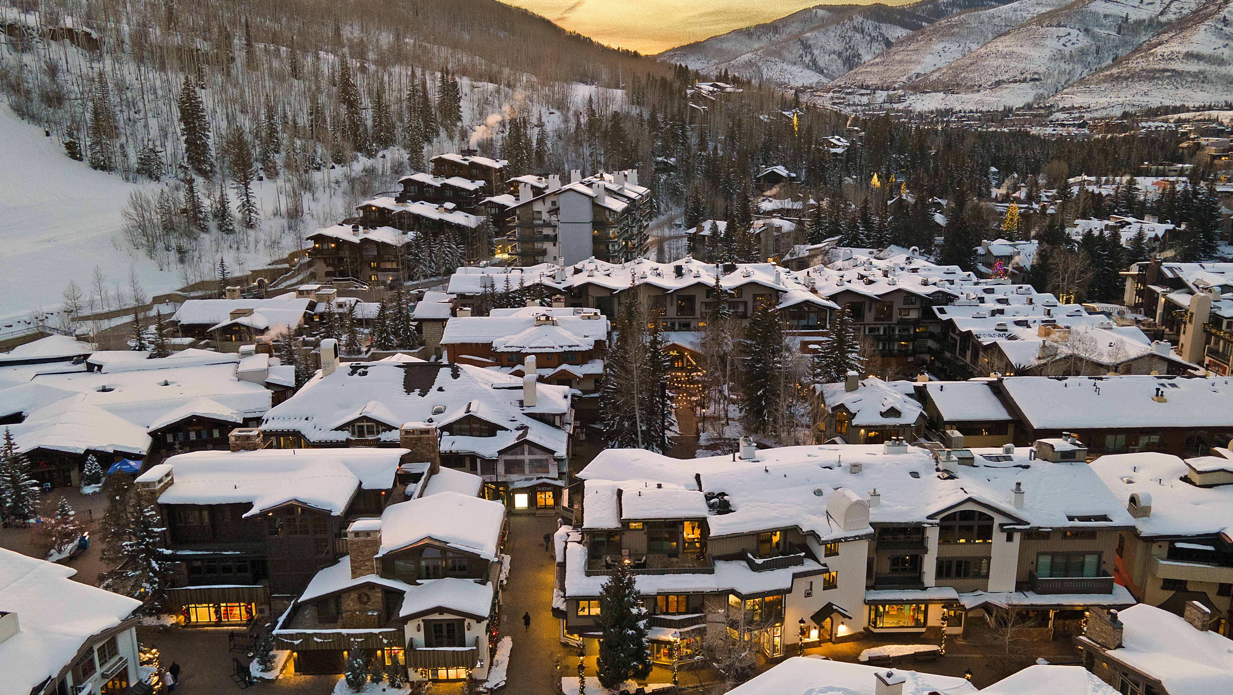 This aerial view showcases a luxurious ski resort town nestled in a snow-covered mountain landscape. The architecture features chalet-style homes and buildings with snow-laden roofs, creating a picturesque winter scene. The warm glow from the windows and streetlights adds a cozy and inviting ambiance to the overall setting.