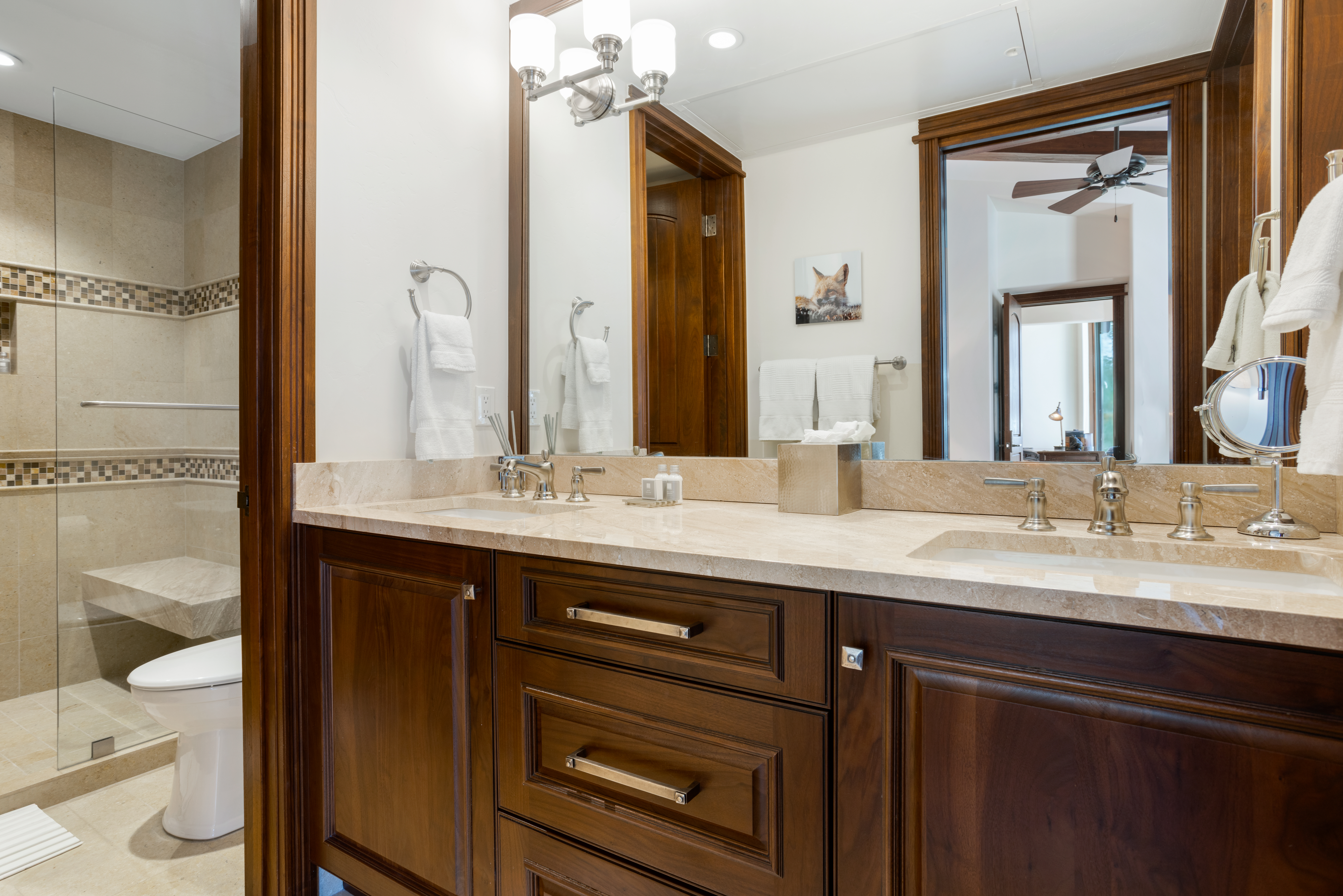 This primary bathroom features a double vanity with a light-colored countertop and dark wood cabinetry. A large mirror reflects the vanity area and a glimpse into another room, while a glass-enclosed shower and toilet are visible to the left. The overall impression is luxurious and well-appointed.