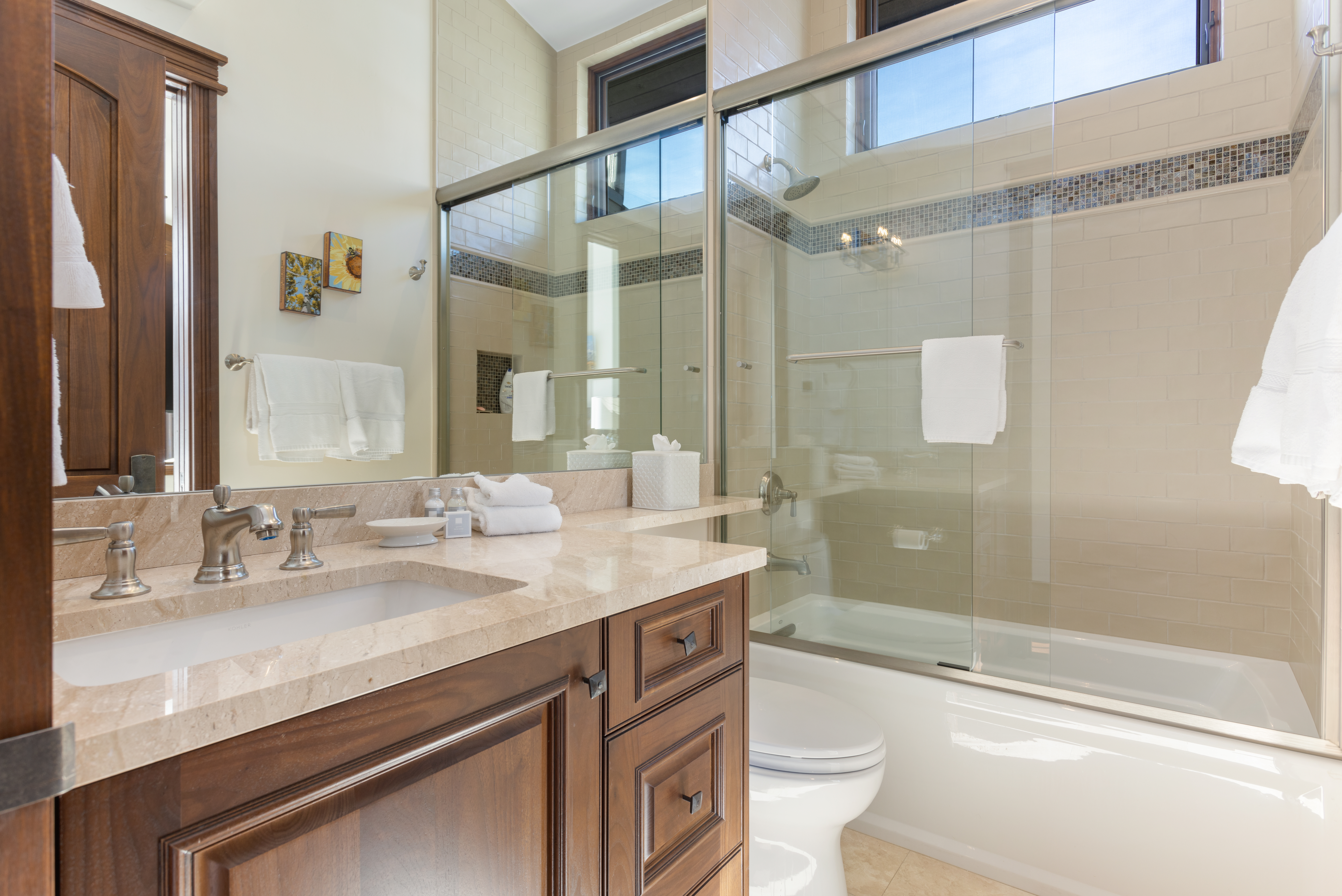 This is a well-lit primary bathroom featuring a vanity with a light-colored stone countertop and dark wood cabinetry. A large mirror reflects the space, including a glass-enclosed shower and bathtub combination with tiled walls and a decorative mosaic accent. The overall impression is clean, modern, and luxurious.