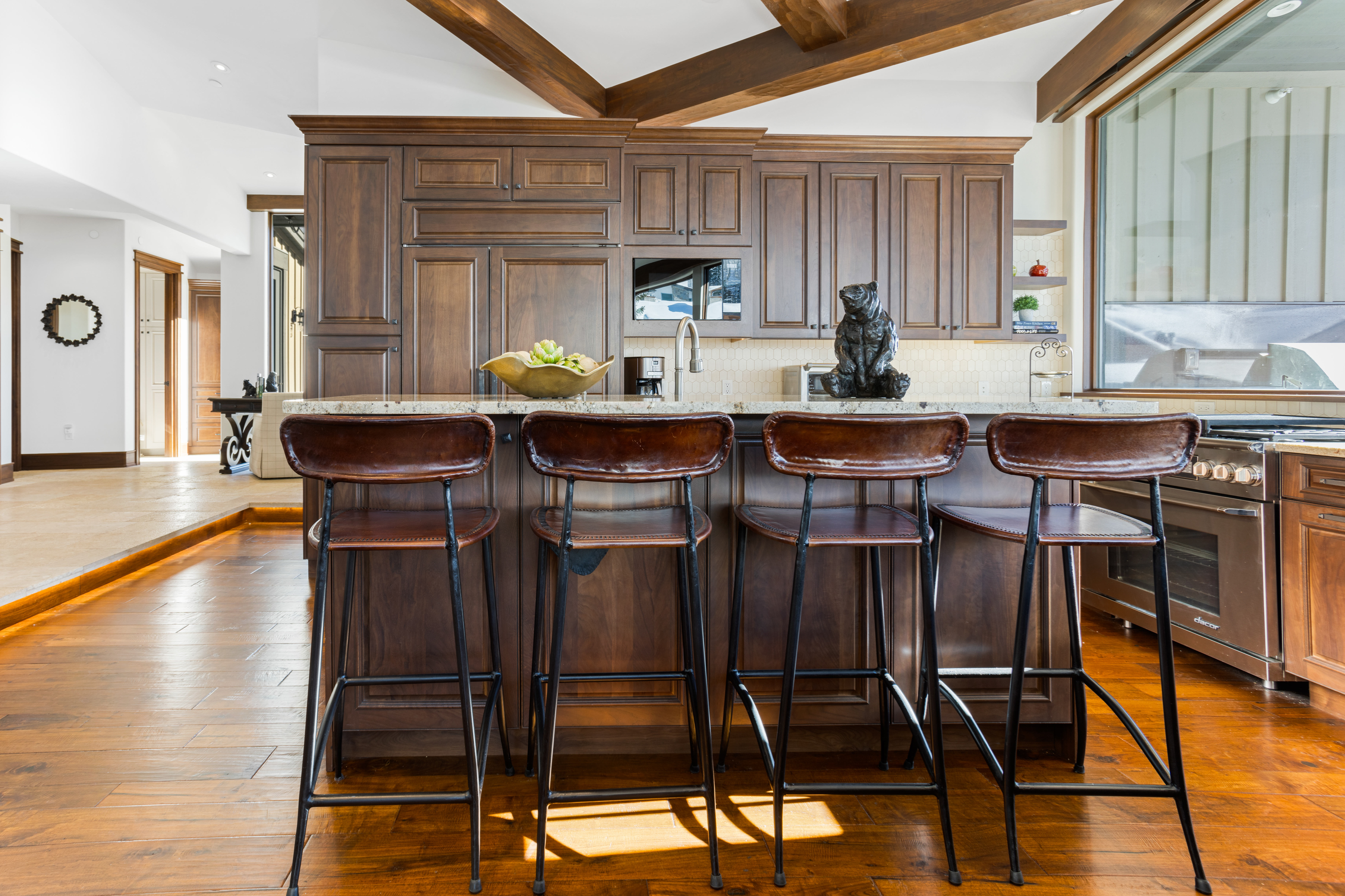 This is a warm and inviting kitchen featuring dark wood cabinetry, granite countertops, and stainless steel appliances. A kitchen island with four leather bar stools provides seating, while wooden beams add character to the ceiling. The hardwood floors and natural light create a cozy atmosphere.