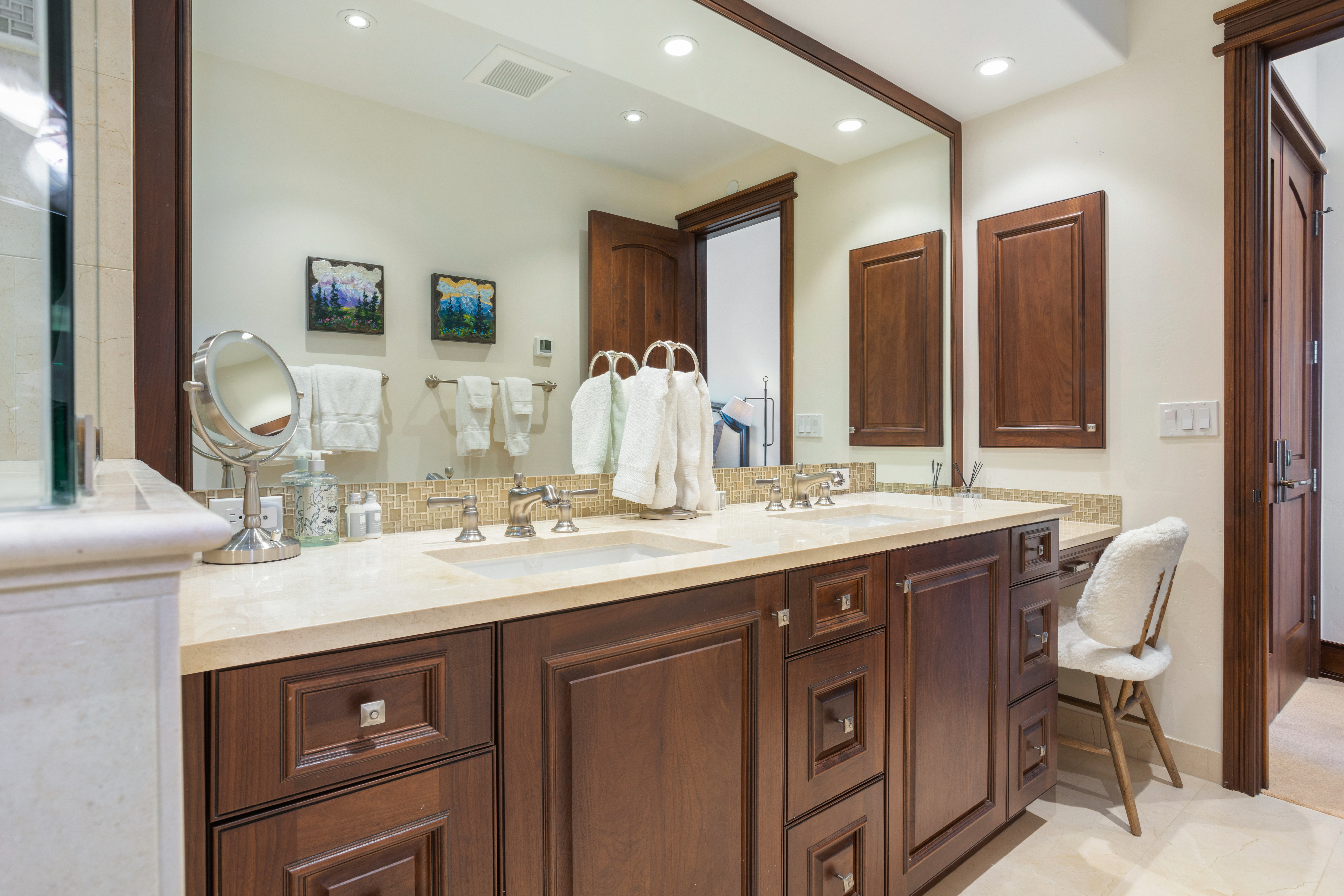 This is a well-lit primary bathroom featuring a double vanity with dark wood cabinetry and a light-colored countertop. A large mirror spans the length of the vanity, reflecting artwork and towels. The bathroom has a luxurious feel with its neutral color palette and elegant fixtures, suggesting a comfortable and upscale space.
