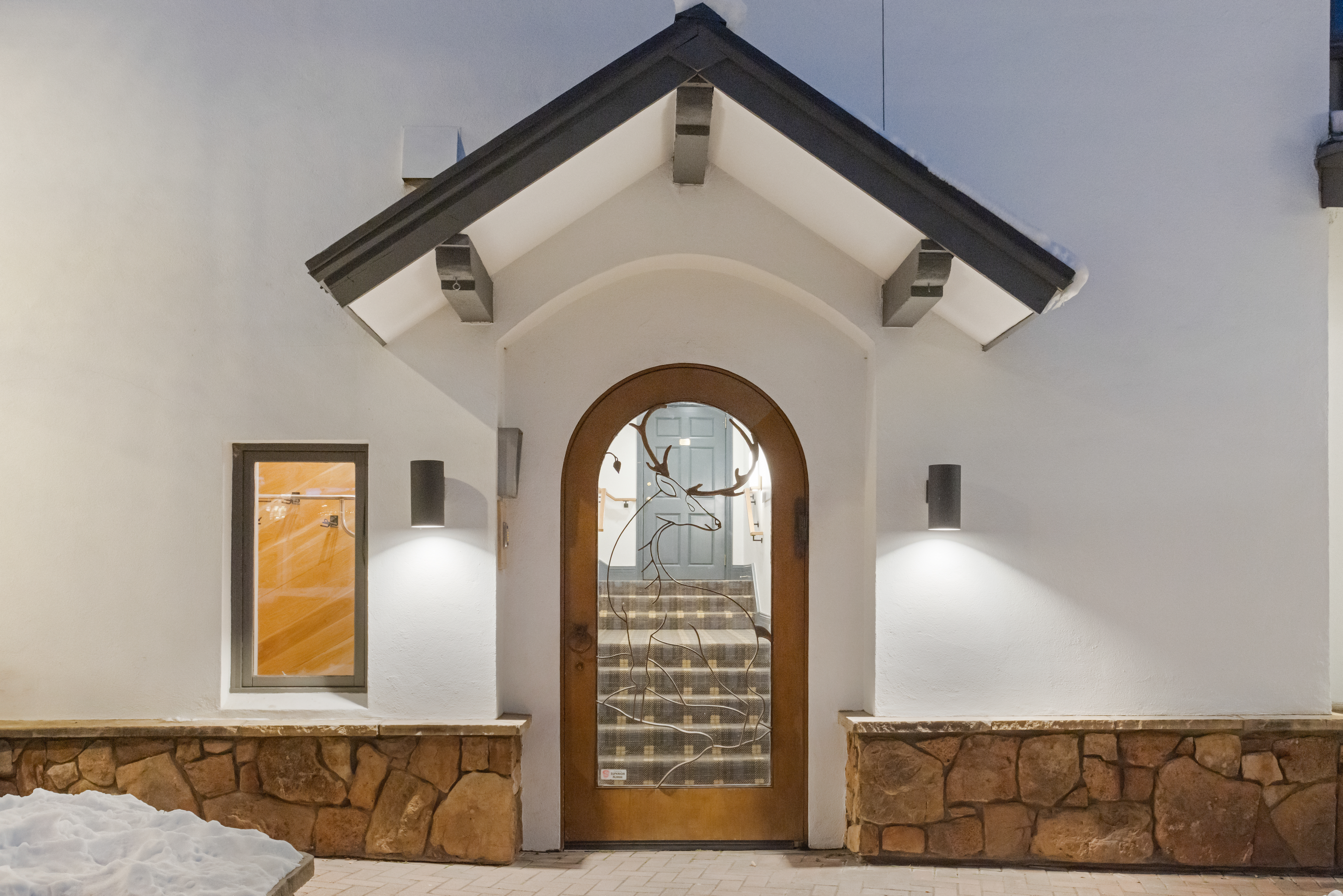 This image showcases an inviting entryway to a home, featuring a unique wooden door with a deer design. The entrance is framed by stone accents and flanked by modern sconce lighting, creating a warm and welcoming ambiance. Above the door is a peaked roof structure with exposed beams, adding architectural interest to the facade.