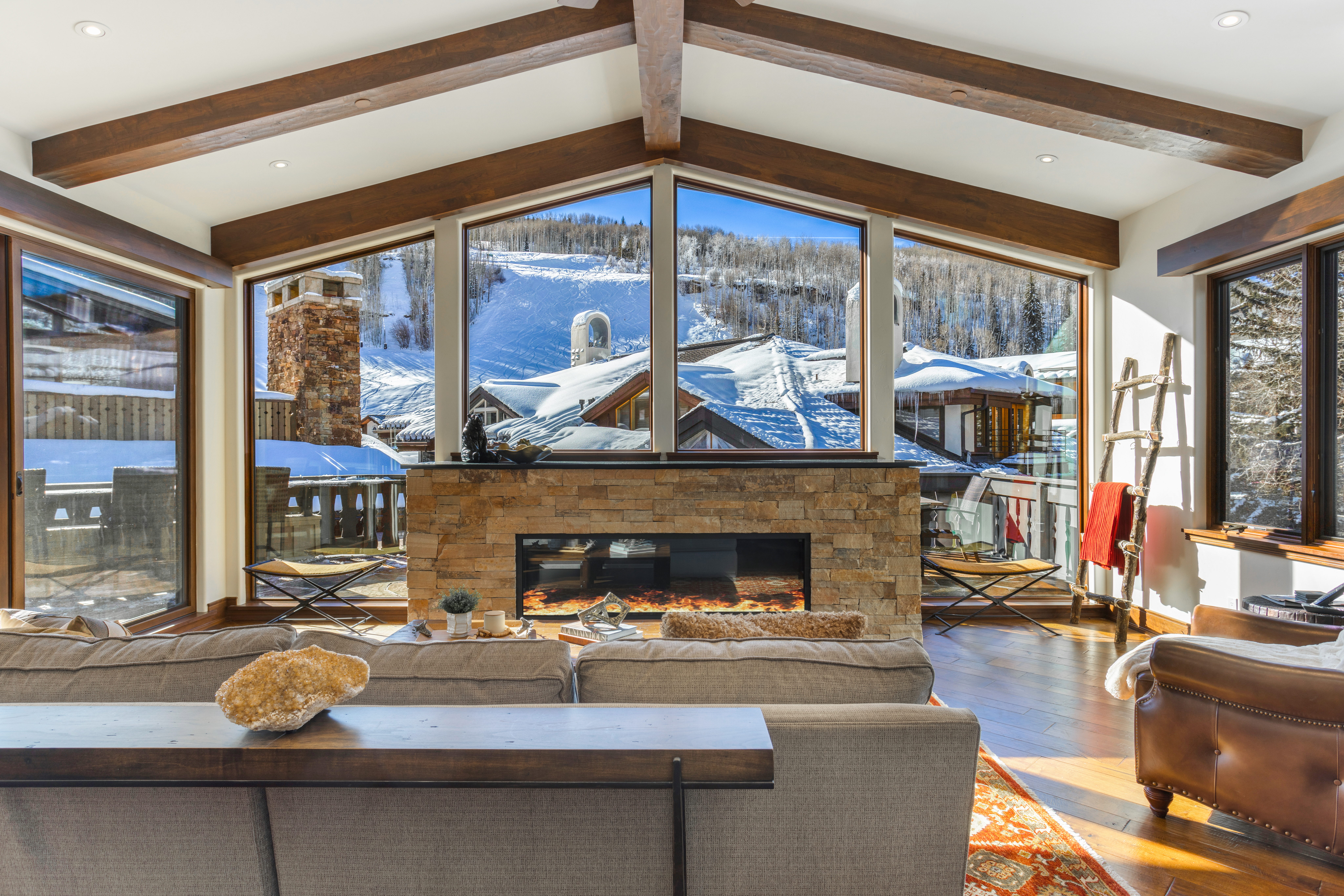 This inviting living room features a stone fireplace with a modern firebox, exposed wooden beams, and large windows offering a picturesque view of a snow-covered landscape. The room is furnished with a comfortable sofa and a leather armchair, creating a cozy and luxurious atmosphere. The hardwood floors and area rug add warmth and texture to the space.