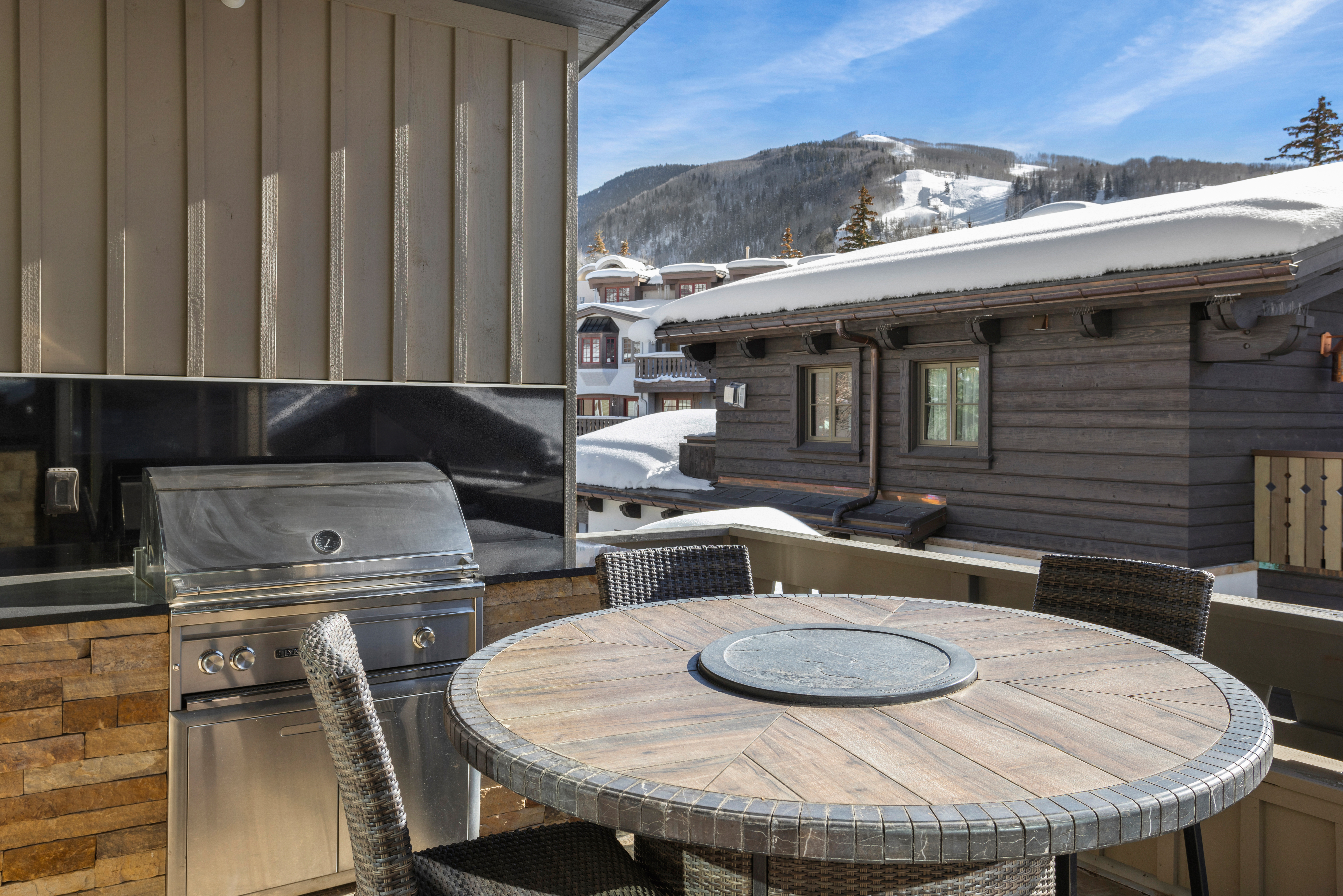 This image showcases a luxurious outdoor patio or balcony area, complete with a built-in grill and a round wooden table surrounded by wicker chairs. The setting offers a scenic mountain view, suggesting a high-end property perfect for entertaining or relaxing outdoors. The presence of snow on the rooftops indicates a winter setting, adding to the cozy and inviting atmosphere.
