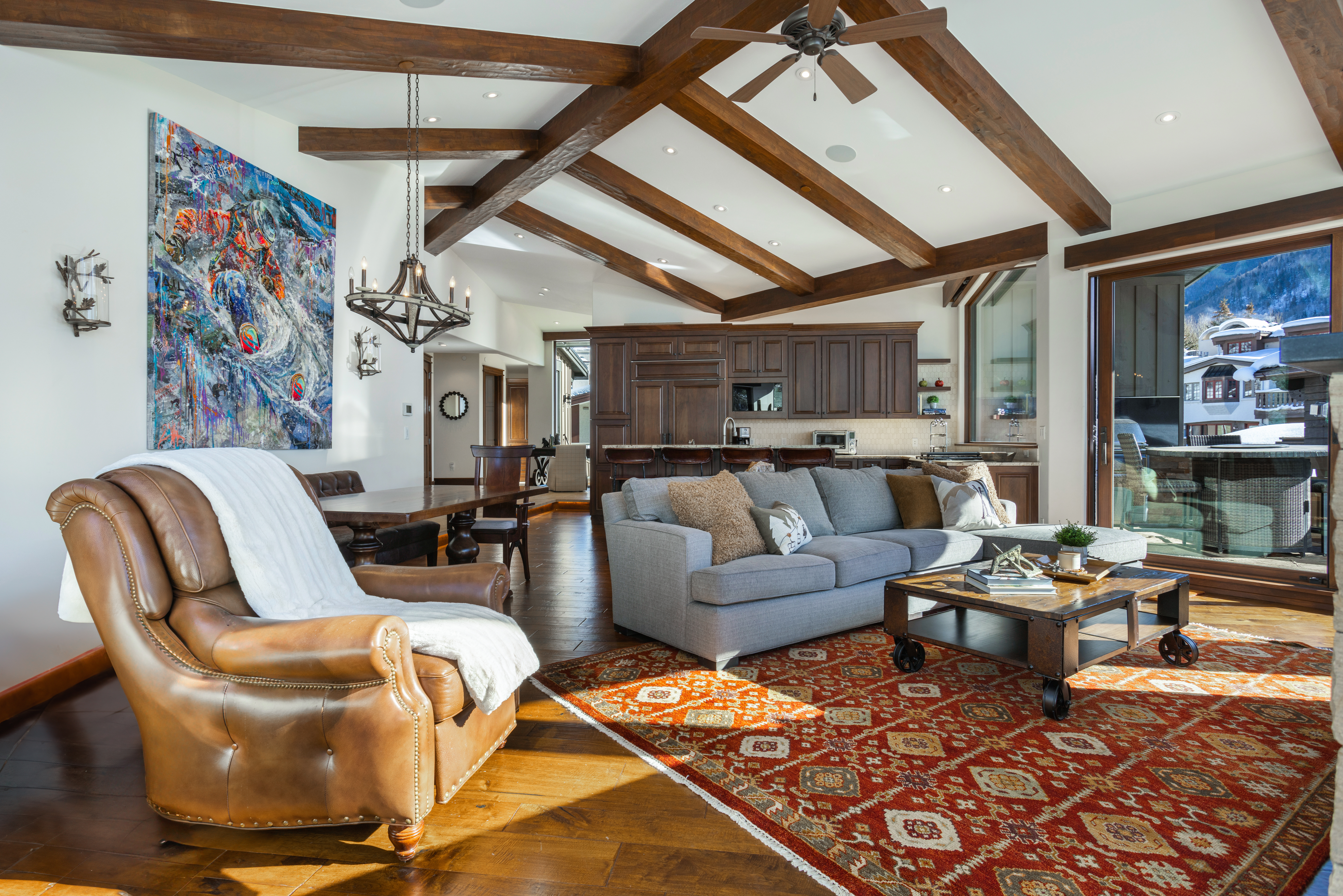 This is an interior shot of a luxurious living room featuring exposed wooden beams on the ceiling, a comfortable gray sofa, and a leather armchair. A vibrant abstract painting adorns the wall, and a rustic coffee table sits on a patterned rug, adding warmth and character to the space. The open layout seamlessly connects the living area to the kitchen, creating an inviting and spacious atmosphere.