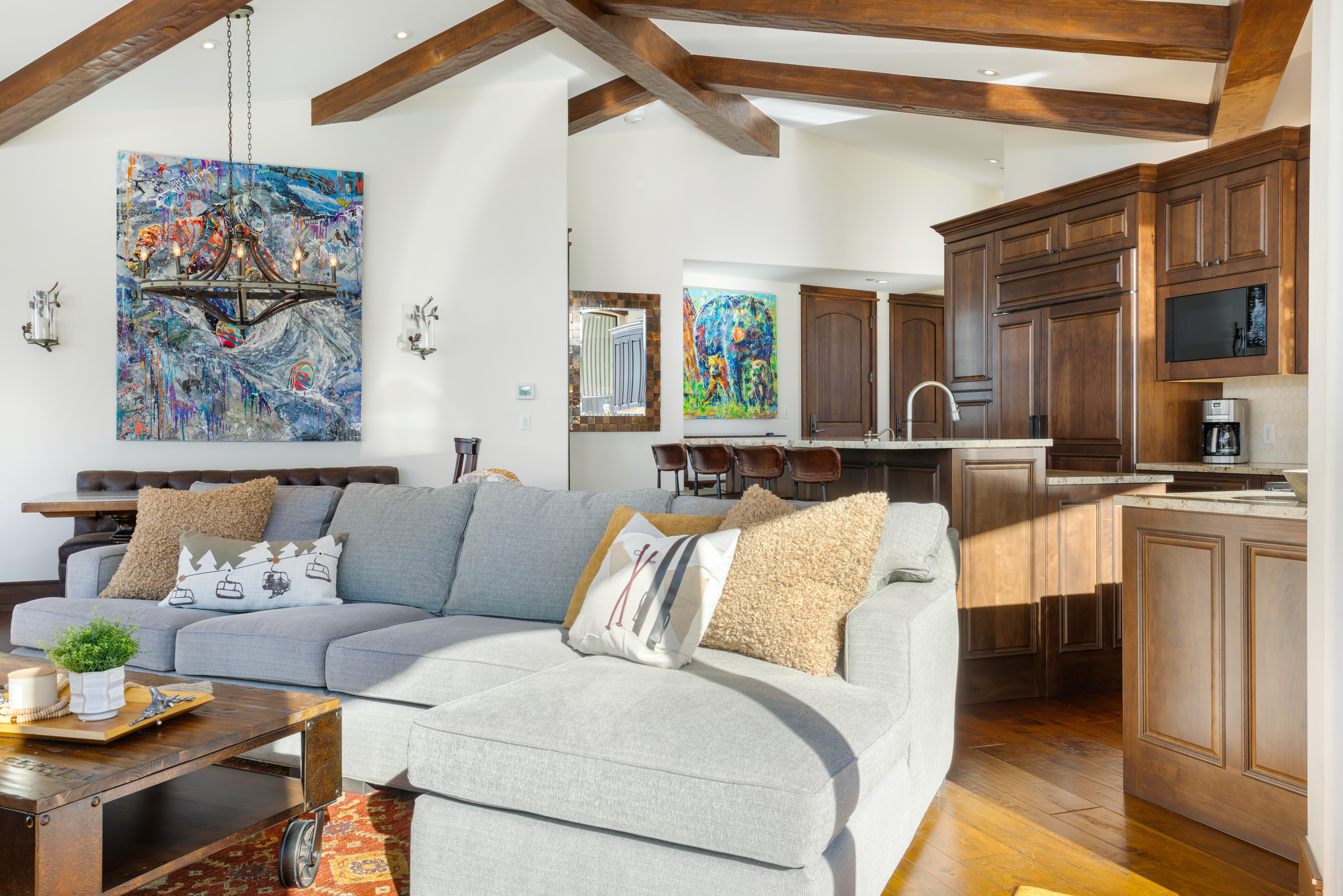 This inviting living room features a large, comfortable gray sectional sofa adorned with decorative pillows, a rustic wooden coffee table, and an eye-catching abstract painting above the sofa. Exposed wooden beams add character to the high ceilings, while the open layout seamlessly connects the living area to the kitchen, creating a warm and welcoming atmosphere. The perspective is from a low angle, emphasizing the depth and spaciousness of the room.
