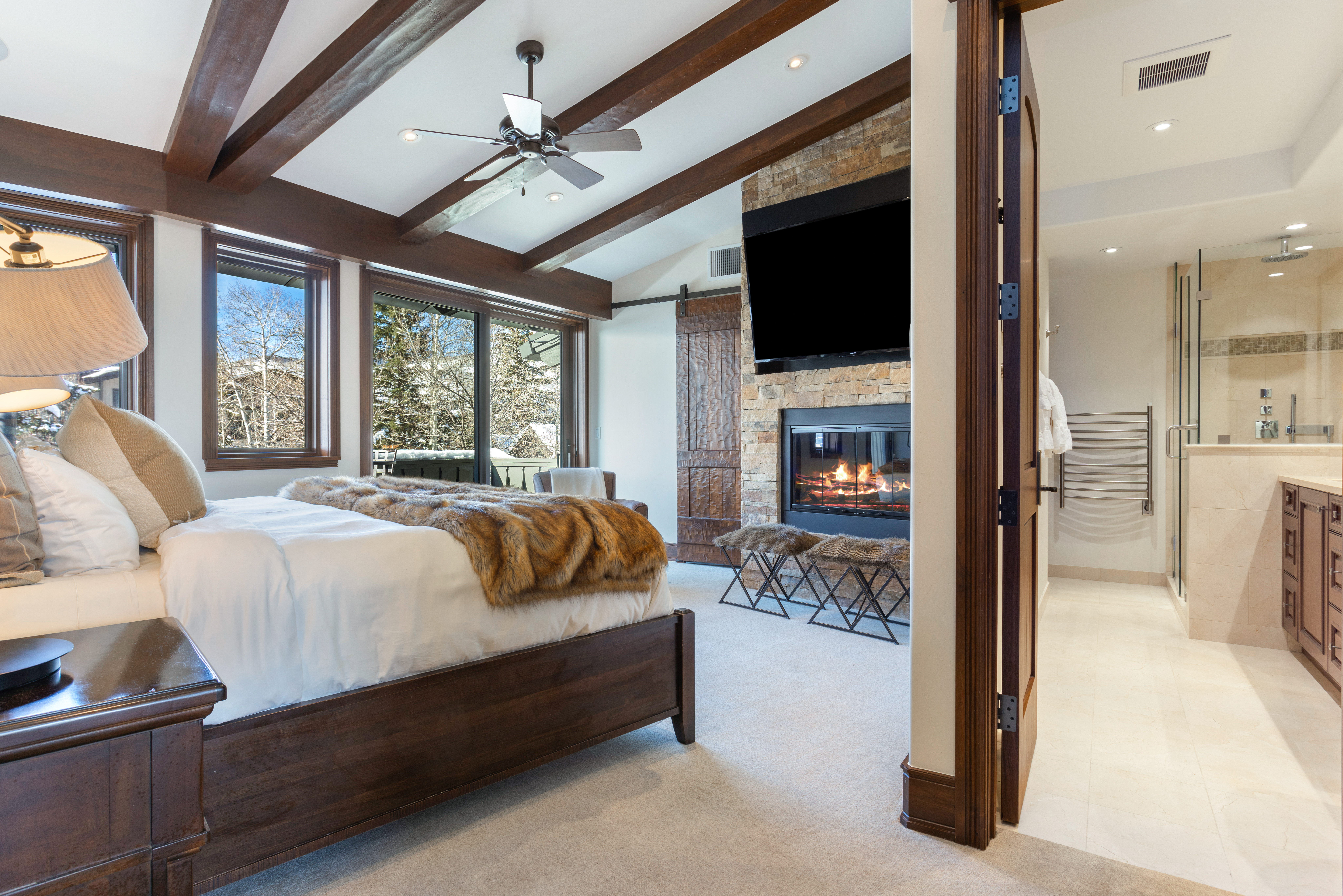 This is a primary bedroom featuring a king-size bed with a fur throw, a stone fireplace with a mounted TV, and exposed wood beams on the ceiling. Large windows offer a view of a snowy landscape, and an open doorway leads to a luxurious bathroom. The room exudes a cozy and upscale atmosphere.