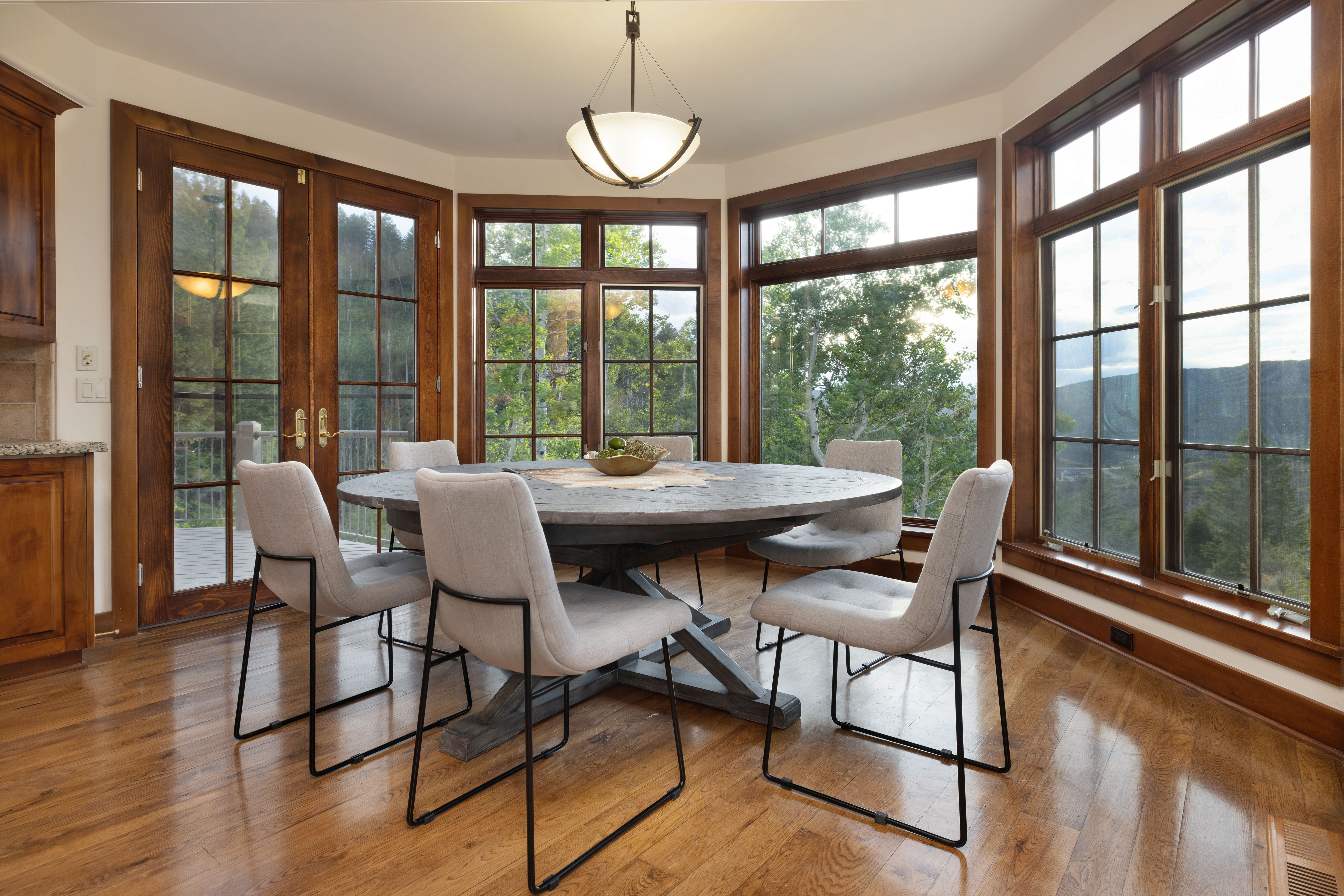 This is a dining room featuring a round wooden table with six upholstered chairs. Large windows surround the dining area, offering views of the surrounding trees and landscape. A modern chandelier hangs above the table, and the hardwood floors add warmth to the space.