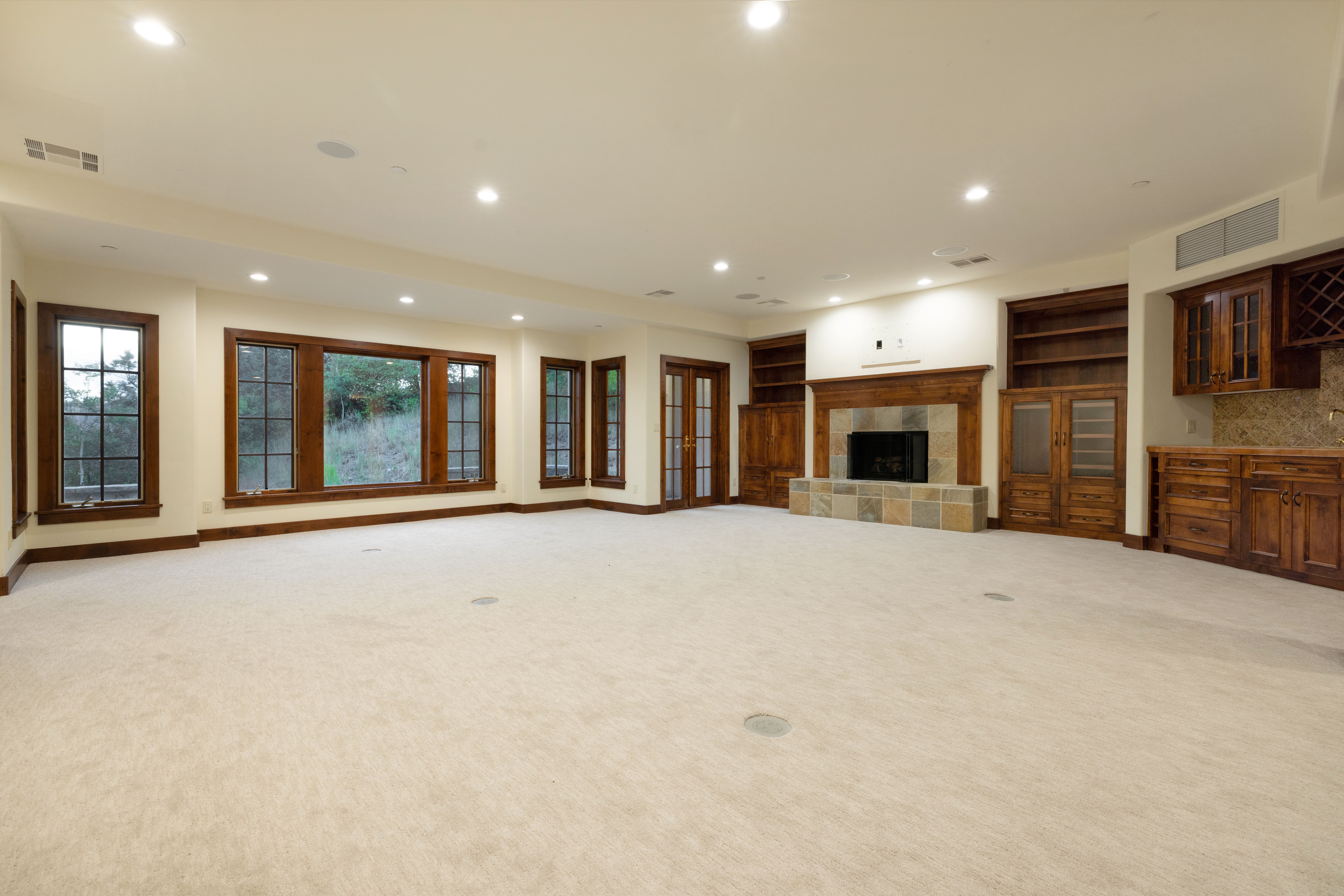 This is a spacious living room featuring neutral-toned carpet, wood-framed windows, and a fireplace with stone accents. Built-in wooden cabinets and shelving flank the fireplace, adding storage and character. The room is well-lit with recessed lighting, creating a warm and inviting atmosphere.