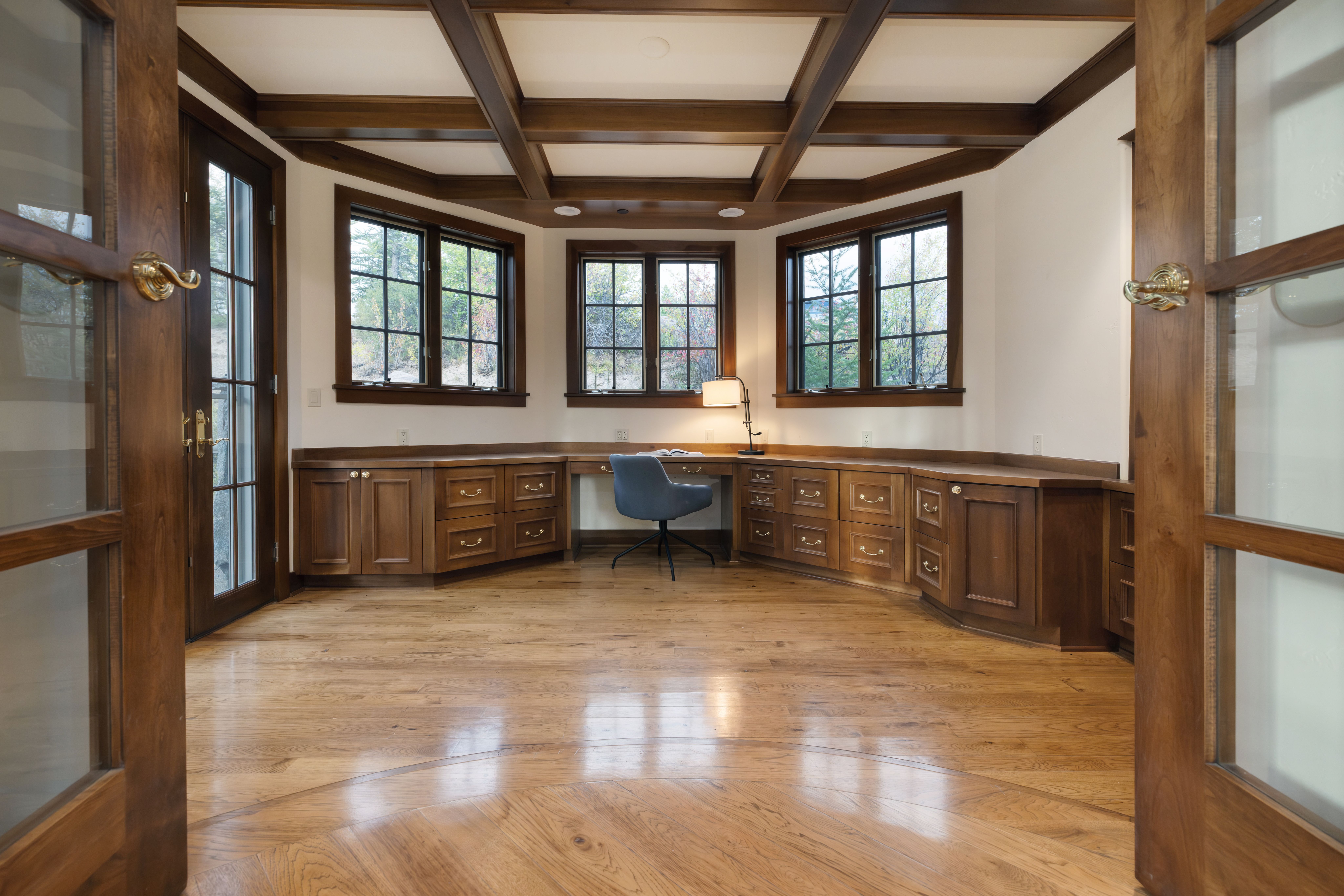 This is an interior shot of a home office or study, featuring custom wood cabinetry and a built-in desk area beneath three windows. The room has a coffered ceiling with wooden beams, and the hardwood flooring adds warmth to the space. The open doorway invites you into the room.