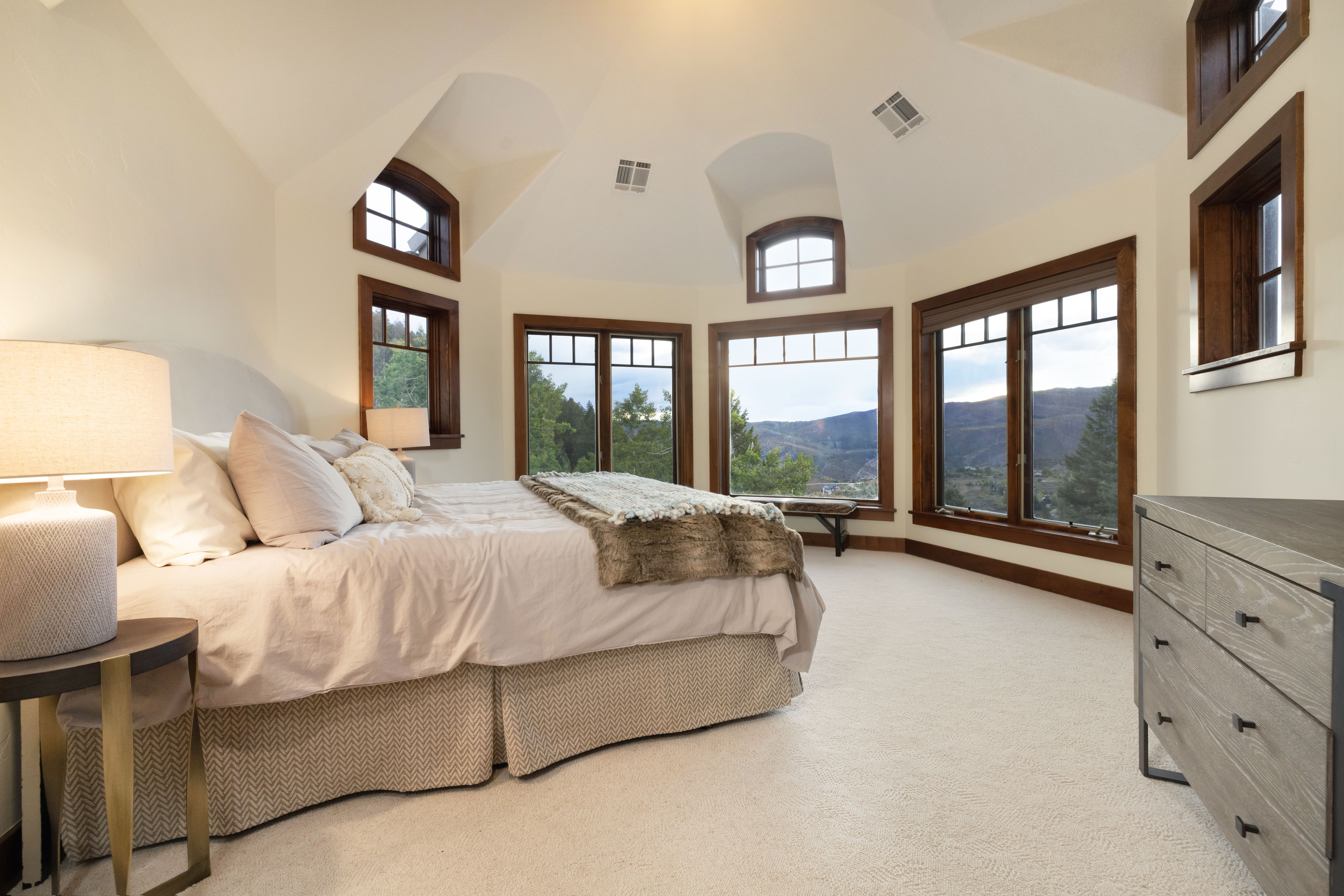 This is a spacious primary bedroom featuring a large bed with neutral-toned linens and a decorative throw. The room is well-lit with natural light streaming through multiple windows offering scenic views. A dresser is visible on the right, and the overall style is elegant and comfortable.
