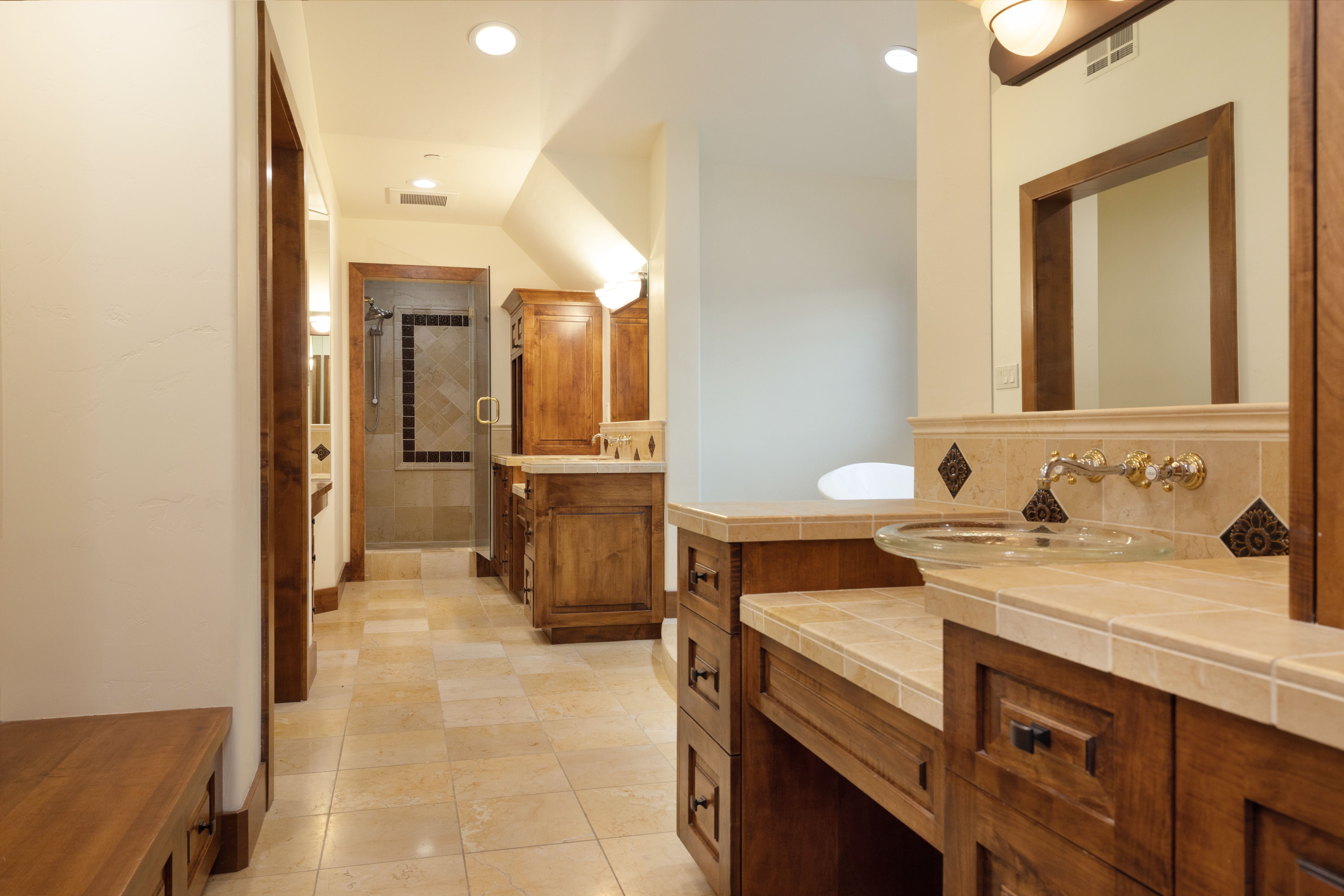 This is a primary bathroom featuring warm-toned tile flooring and wooden cabinetry. The vanity has a glass vessel sink and decorative tile accents. A glass-enclosed shower is visible in the background, contributing to the spa-like atmosphere of the space.