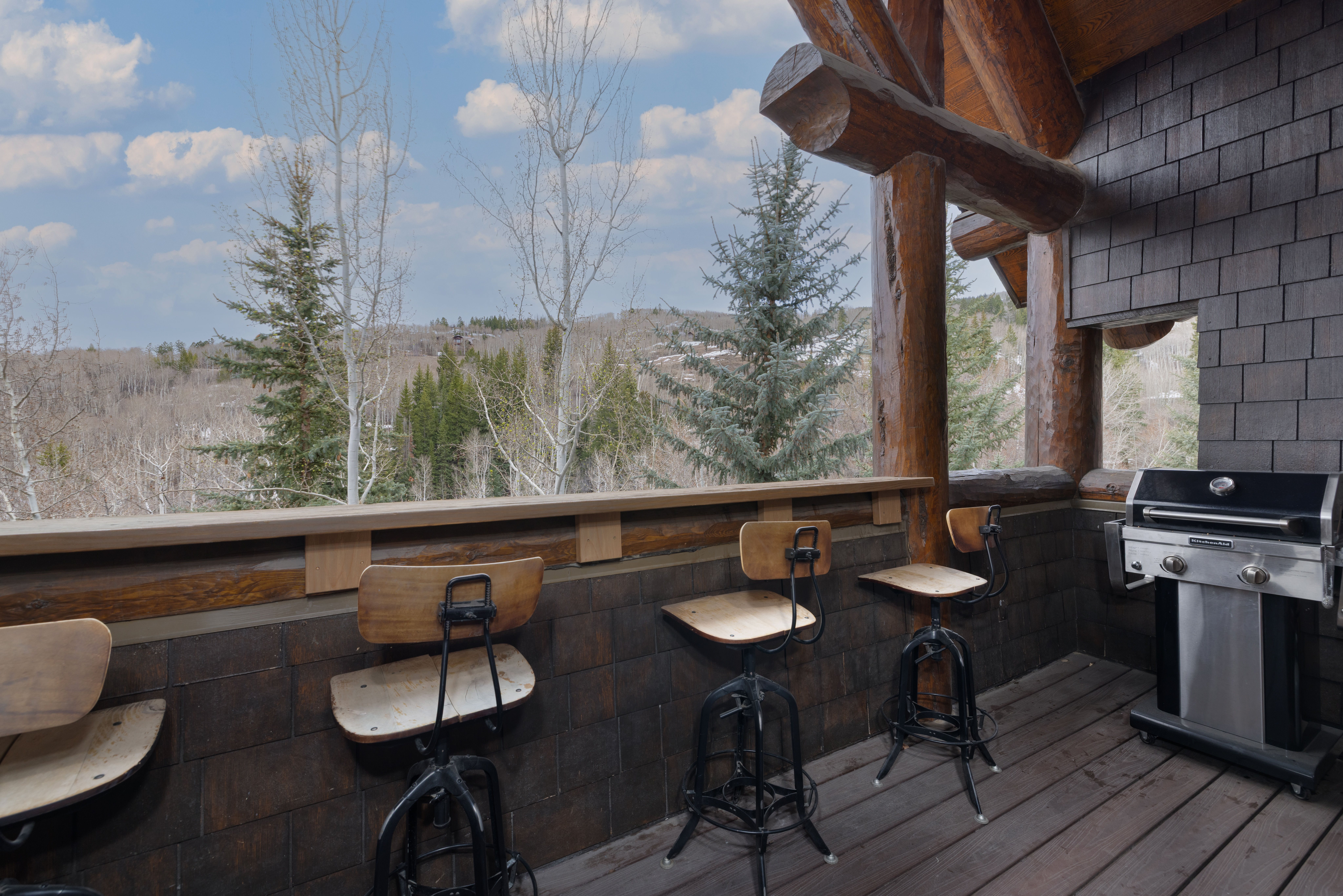 This image showcases a rustic outdoor patio or balcony area, featuring wooden decking and log beam construction. The space includes bar-style seating and a built-in grill, suggesting an area designed for outdoor cooking and relaxation. The backdrop reveals a scenic view of trees and hills, enhancing the property's appeal.