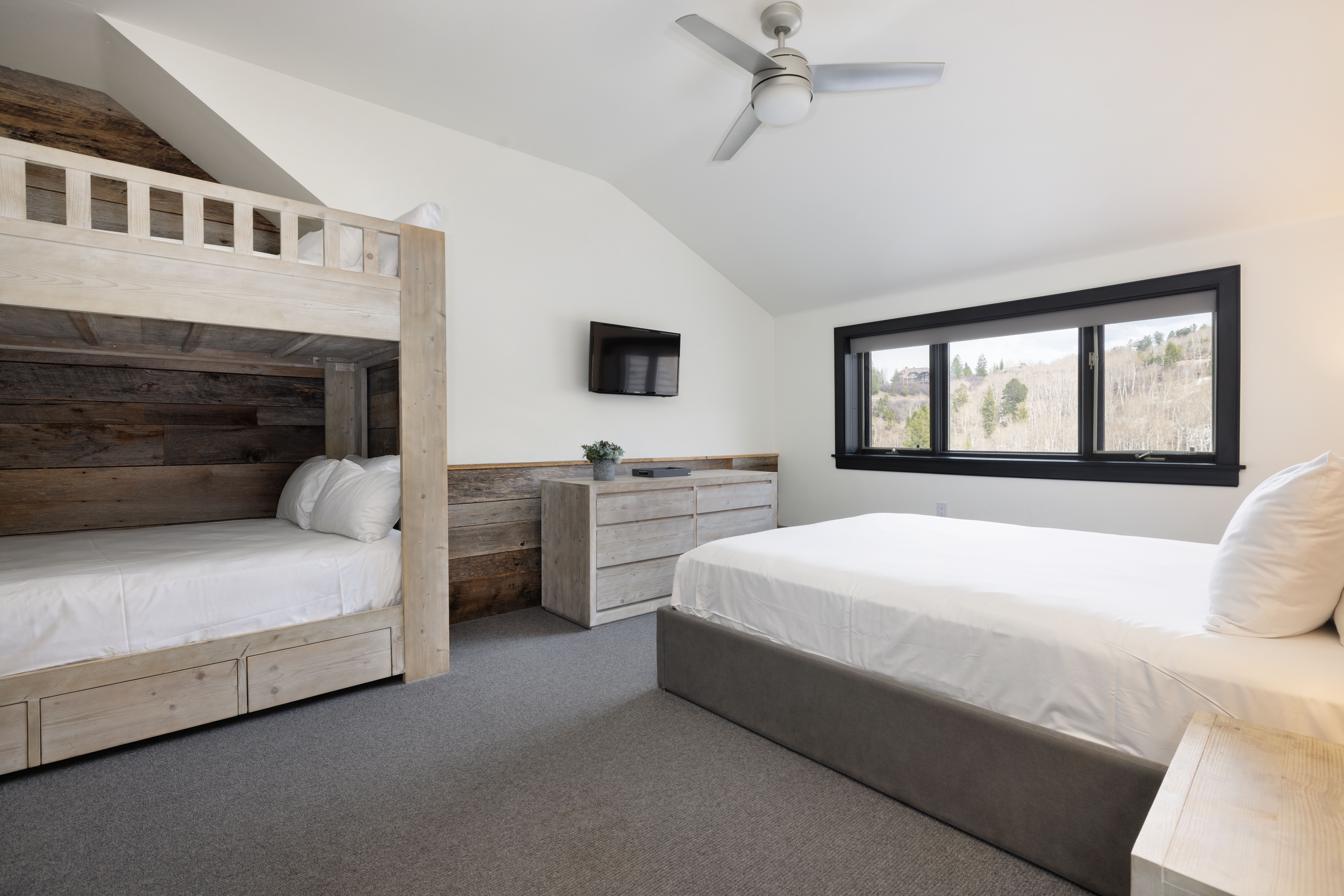 This is a well-lit guest bedroom featuring a bunk bed with a rustic wood accent wall, a queen-sized bed, and a dresser with a television mounted above. The room has a neutral color palette with white walls and gray carpeting, creating a calm and inviting atmosphere. A large window provides natural light and a view of the surrounding landscape.
