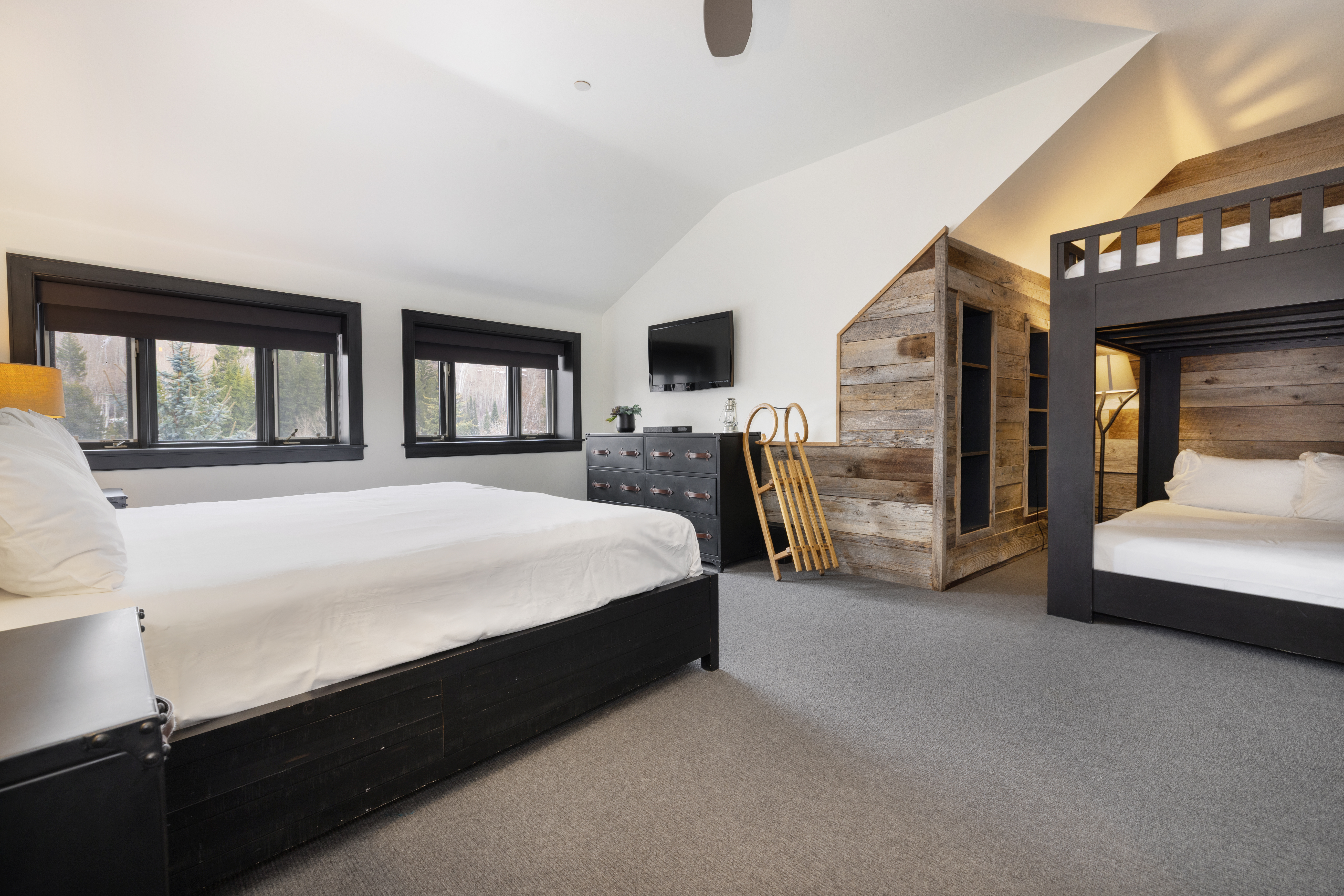 This is a cozy guest bedroom featuring a black bed frame with a white mattress and bedding, complemented by a dark wood dresser and a built-in bunk bed area with rustic wood paneling. The room has two windows with black frames, allowing natural light to illuminate the space, and the overall design creates a warm and inviting atmosphere.