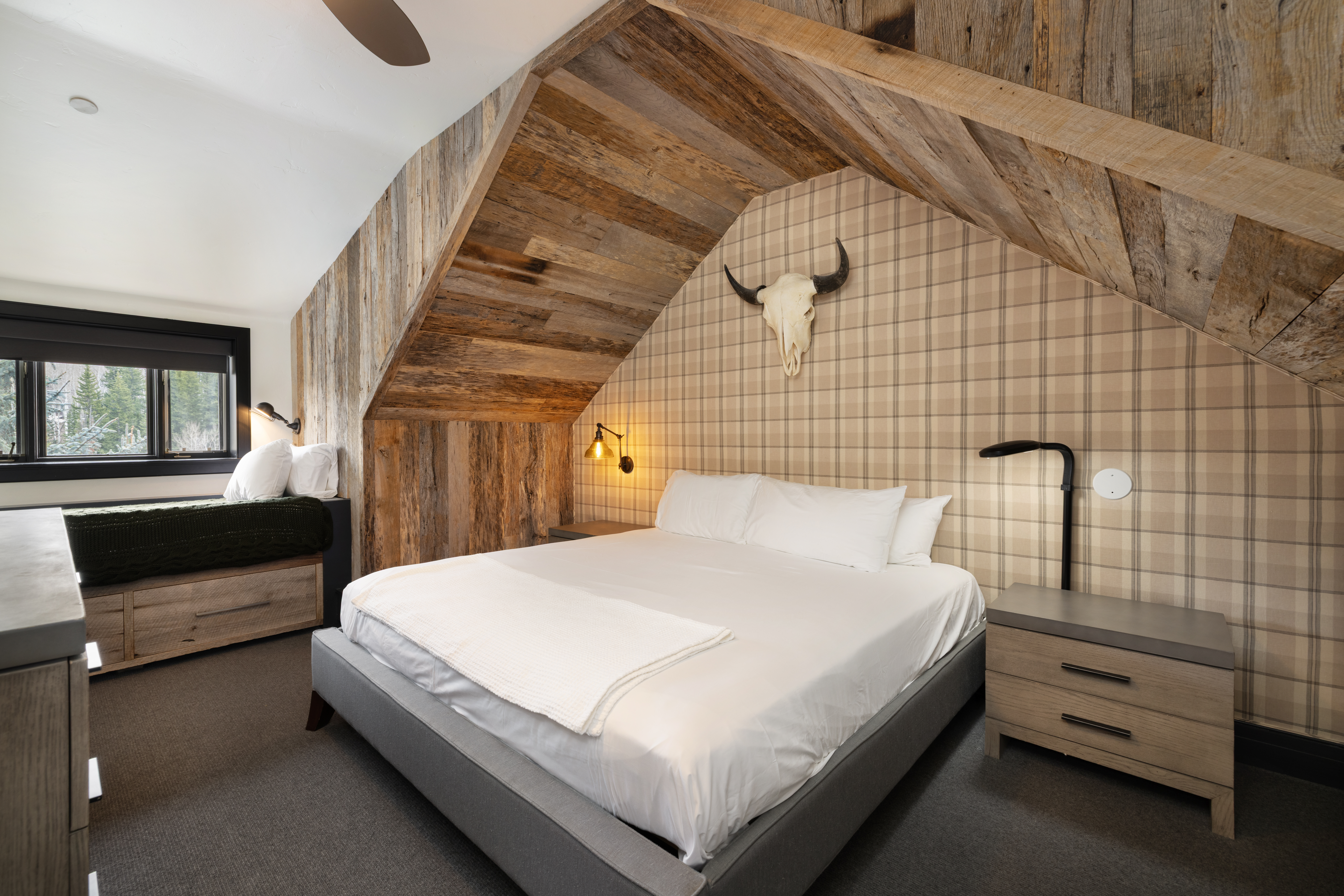 This is a cozy bedroom featuring a bed with white linens, flanked by a nightstand with a lamp. The room has a rustic-chic design with wood paneling on the ceiling and one wall, complemented by plaid wallpaper on another wall. A decorative bull skull adds a unique touch above the bed, and a window seat provides additional seating.