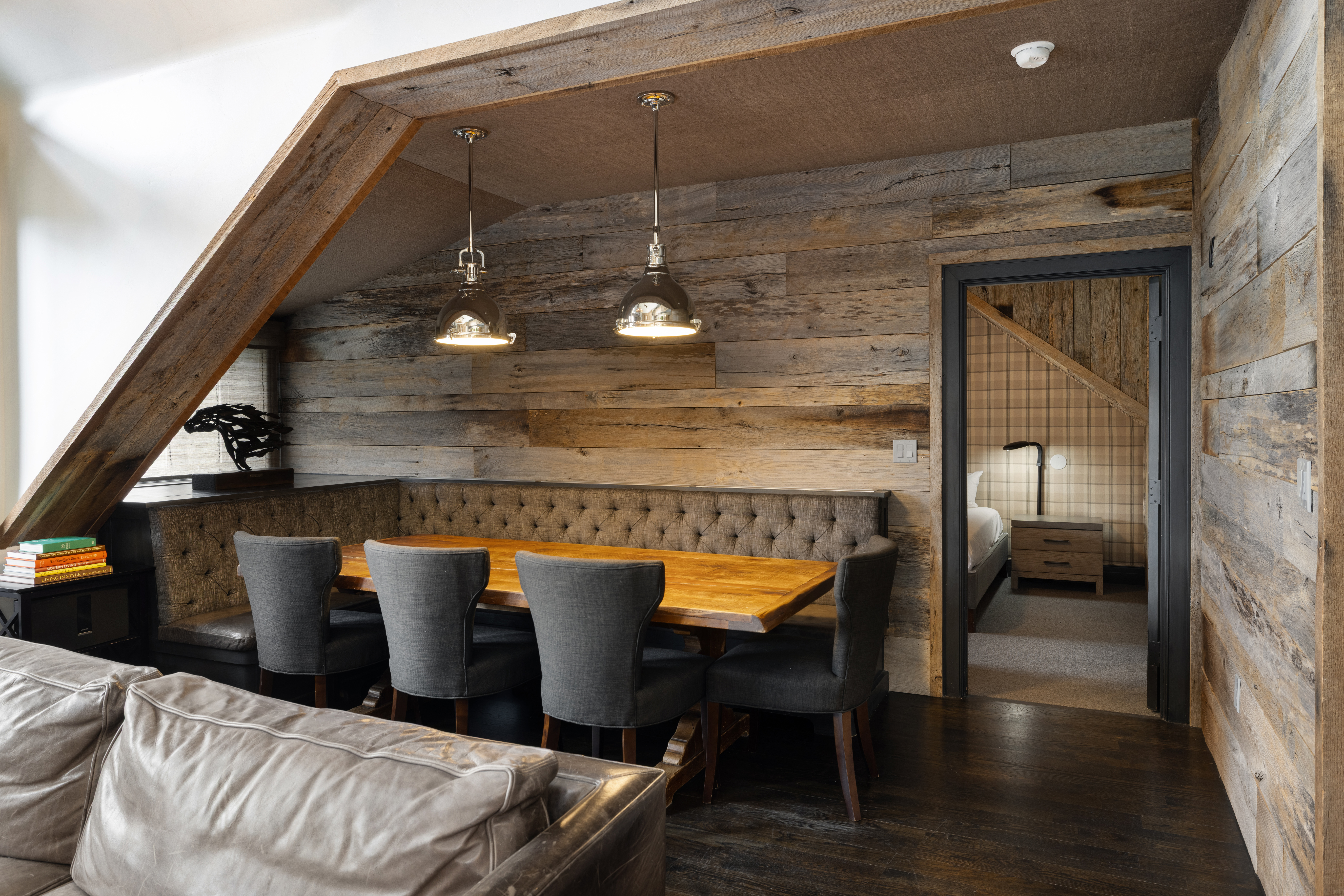 This interior shot showcases a rustic dining area with a built-in upholstered bench and four gray chairs around a wooden table. The walls are clad in reclaimed wood, complemented by industrial-style pendant lighting. An open doorway leads to a bedroom, adding depth and suggesting a cozy, connected living space.