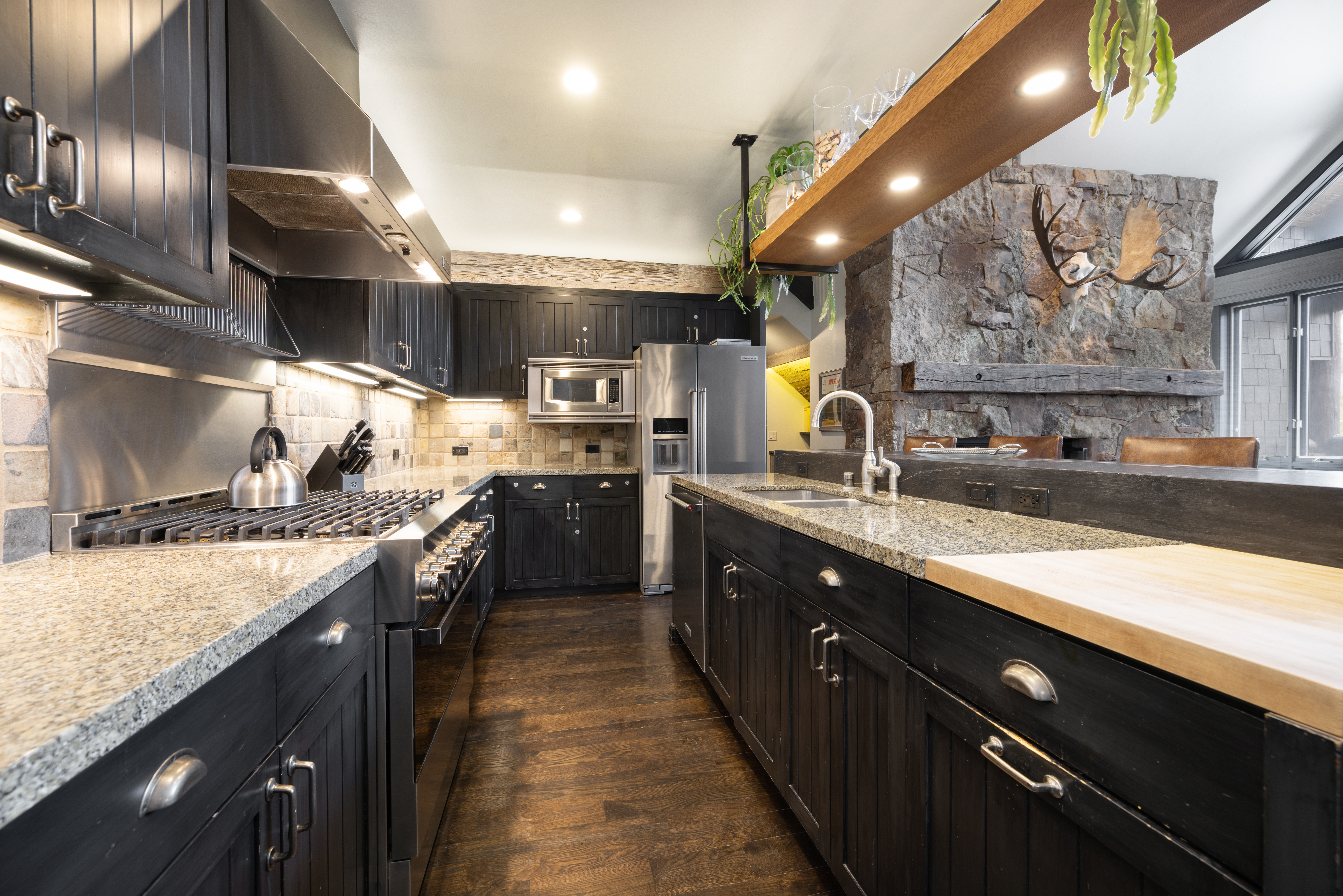 This is a well-appointed kitchen featuring dark wood cabinetry, granite countertops, and stainless steel appliances. The kitchen includes a large range with a stainless steel hood, a built-in microwave, and a stainless steel refrigerator. A stone fireplace is visible in the background, adding a rustic touch to the space.