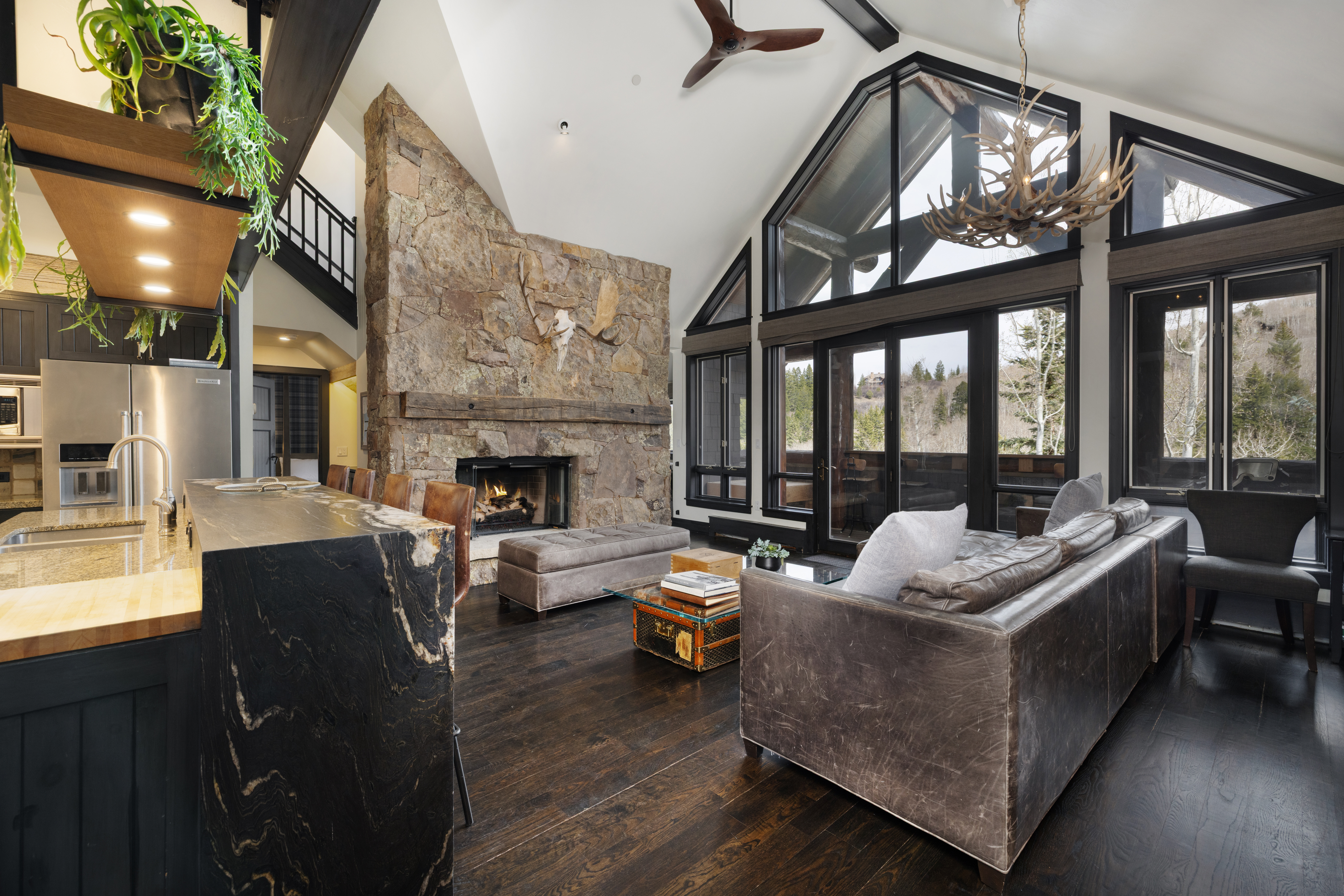 This is an interior shot of a luxurious living room featuring a stone fireplace, a large leather sofa, and floor-to-ceiling windows offering a view of the outdoors. The room has dark hardwood floors and a high, vaulted ceiling with exposed beams, creating a rustic yet elegant atmosphere. A kitchen island is visible in the foreground, suggesting an open-concept design.