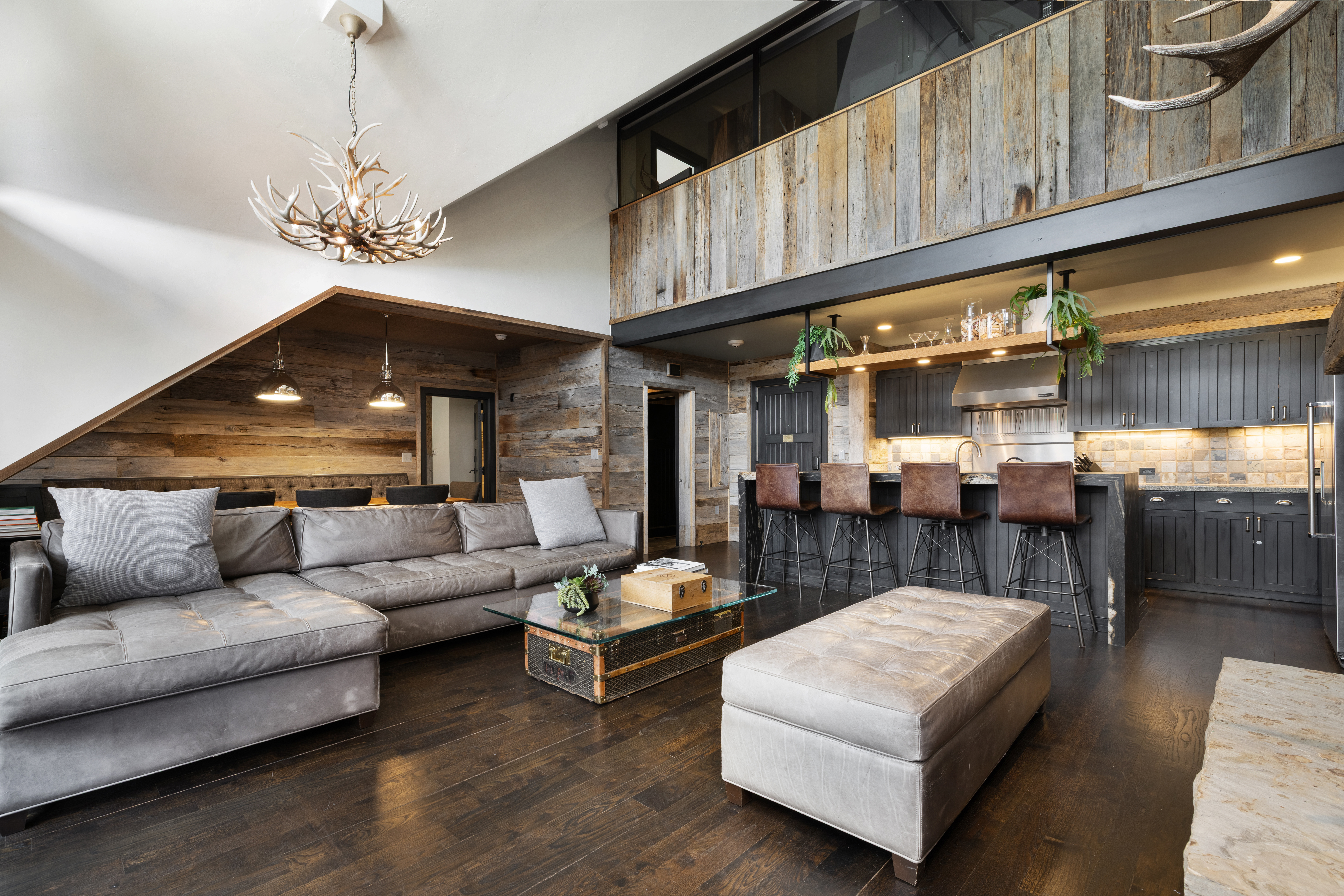 This is an interior shot of a luxurious living room that seamlessly blends with a modern kitchen. The living room features a large, comfortable gray sectional sofa, a unique antler chandelier, and rustic wood paneling. The kitchen boasts dark cabinetry, a breakfast bar with leather stools, and stylish pendant lighting, creating a warm and inviting atmosphere.