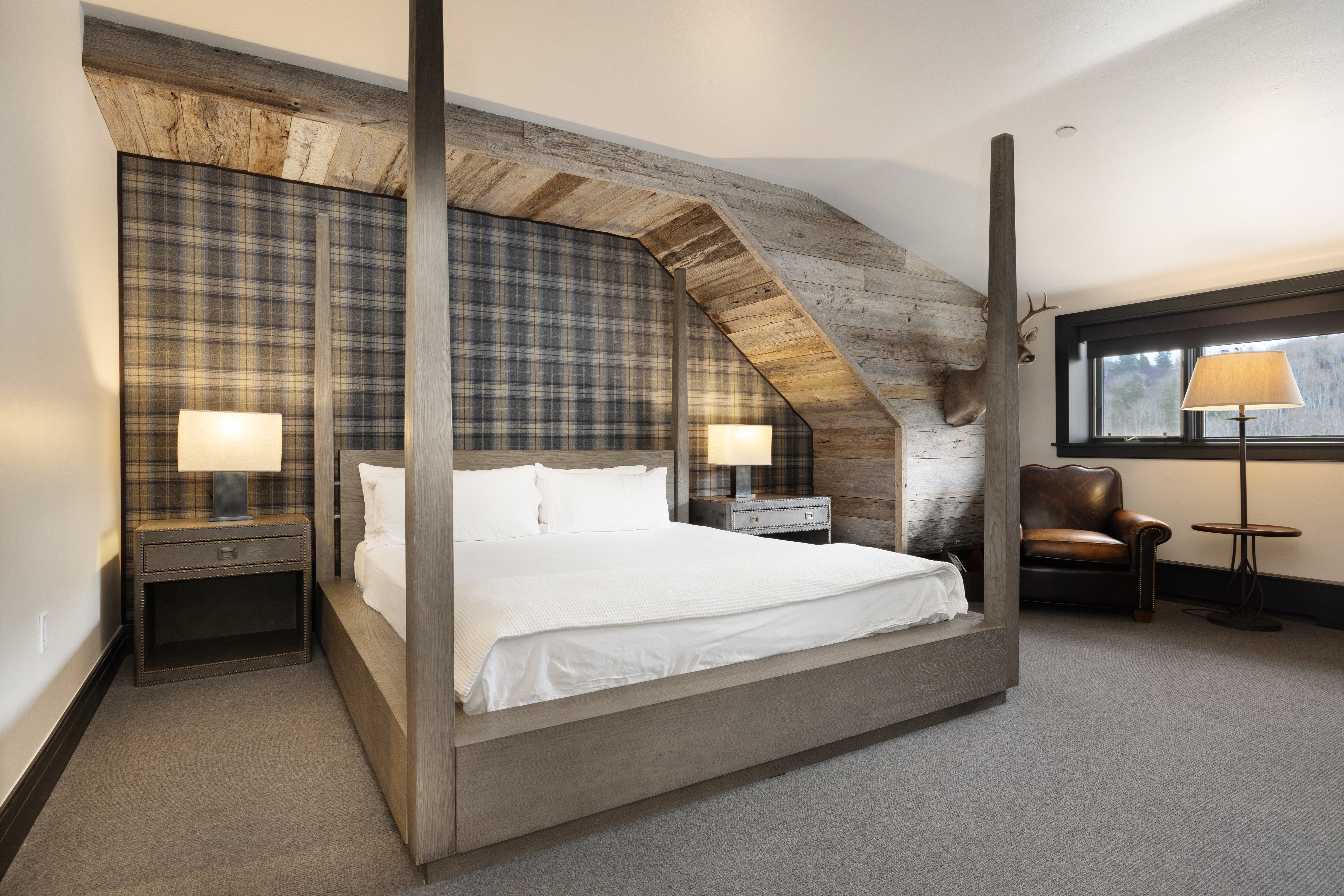 This is a cozy primary bedroom featuring a four-poster bed with white linens, flanked by matching nightstands and lamps. The room has a rustic-chic aesthetic with a plaid accent wall and exposed wooden beams. A leather armchair sits near a window, adding a touch of comfort and sophistication.