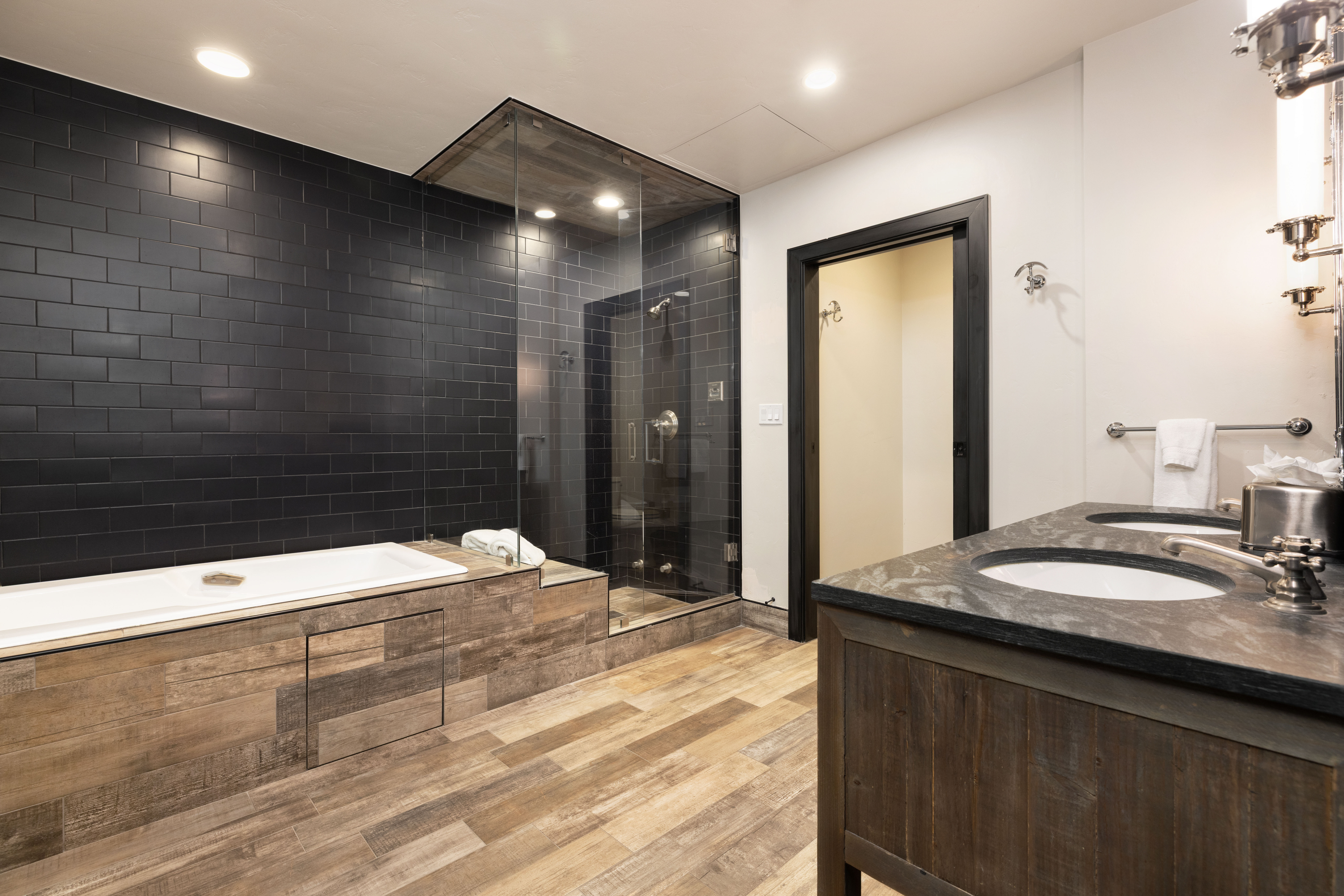 This is a primary bathroom featuring a modern design with a black tiled shower and bathtub area enclosed by glass. The vanity has a dark countertop with dual sinks and rustic wood cabinetry. The flooring is light-colored wood-look planks, and the overall impression is luxurious and contemporary.