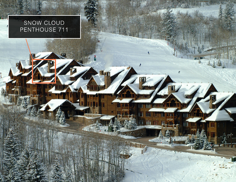 This image showcases the front view of a large, luxurious mountain lodge or condominium complex covered in snow. The architecture features a rustic style with wood and stone elements, multiple gabled roofs, and numerous windows. The setting is a snowy landscape with trees and a ski slope in the background, creating a picturesque winter scene.