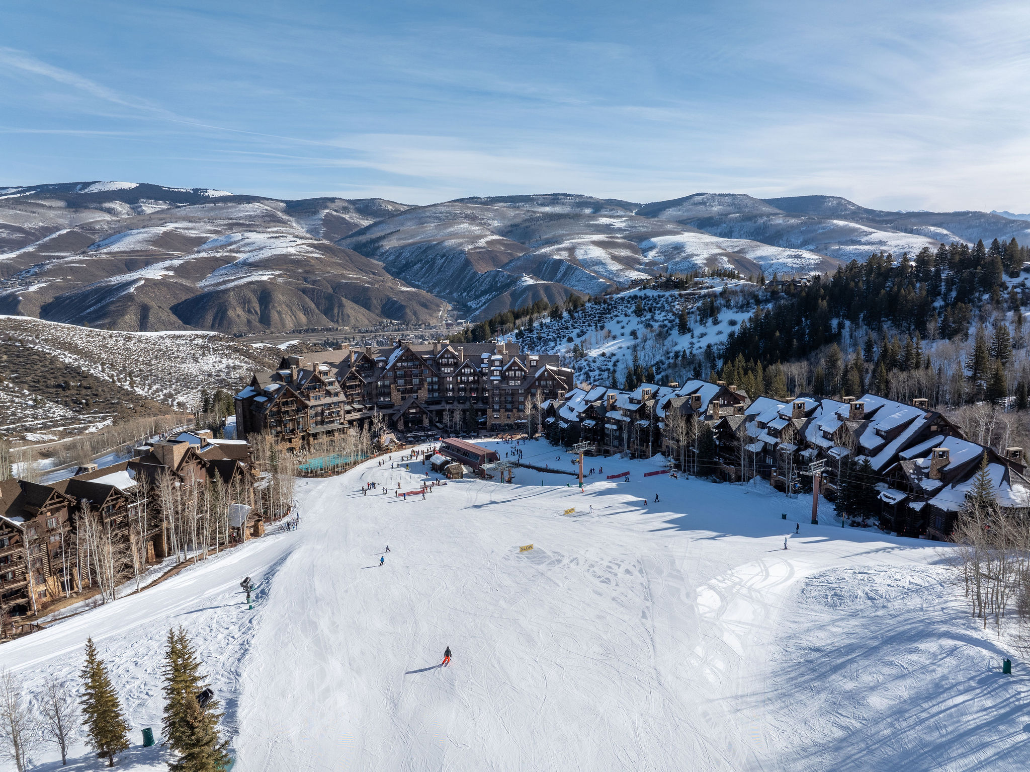 This aerial view showcases a luxurious ski resort nestled in a mountainous landscape. The architecture features chalet-style buildings with dark wood and snow-covered roofs, blending seamlessly with the surrounding evergreen trees. A pristine ski slope dominates the foreground, inviting potential buyers to envision a winter wonderland experience.