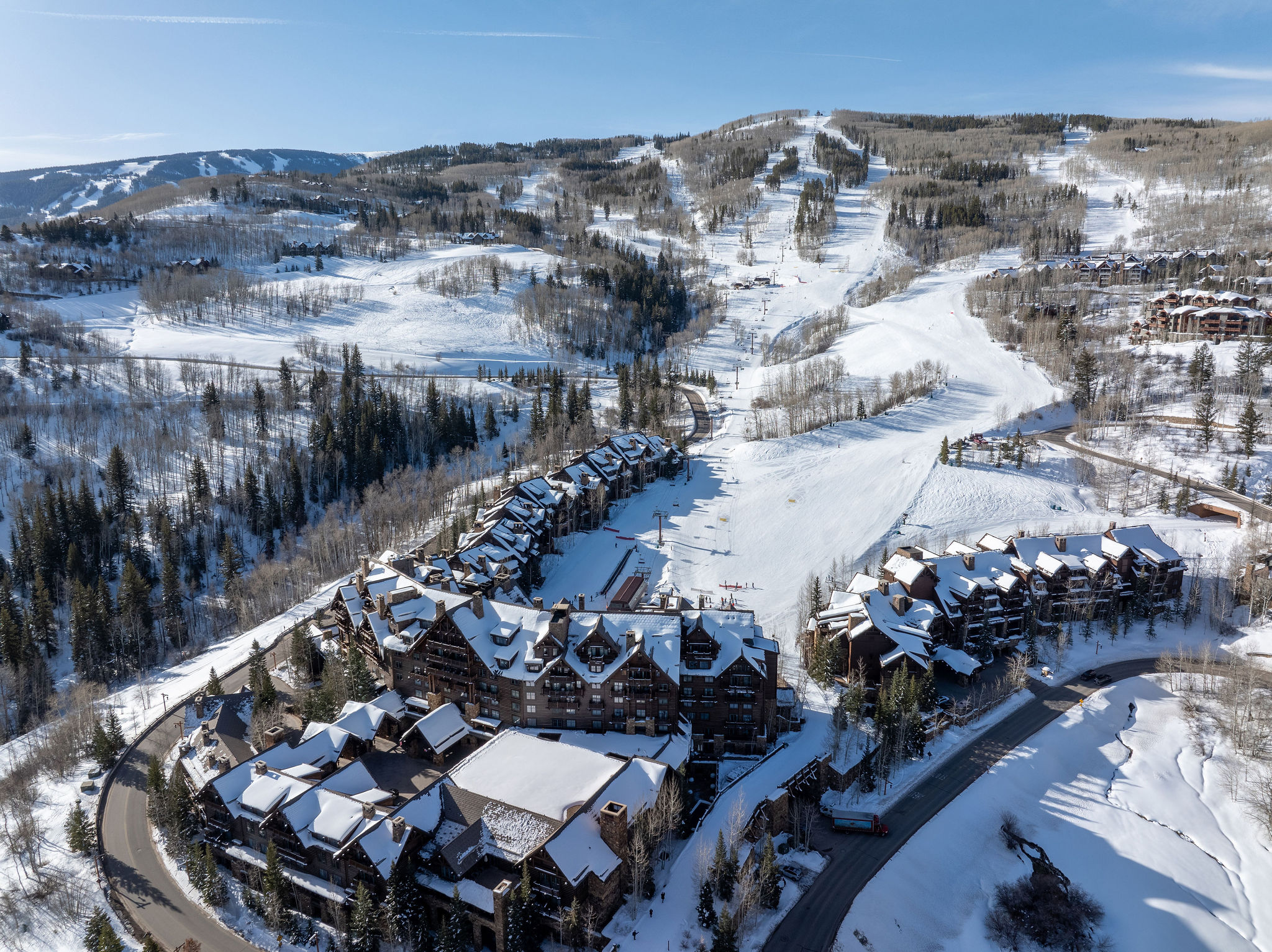 This aerial view showcases a luxurious ski resort nestled in a snow-covered mountain landscape. The buildings feature dark wood exteriors and snow-covered roofs, blending harmoniously with the surrounding evergreen trees and ski slopes. A winding road leads to the resort, suggesting easy access and a sense of seclusion.