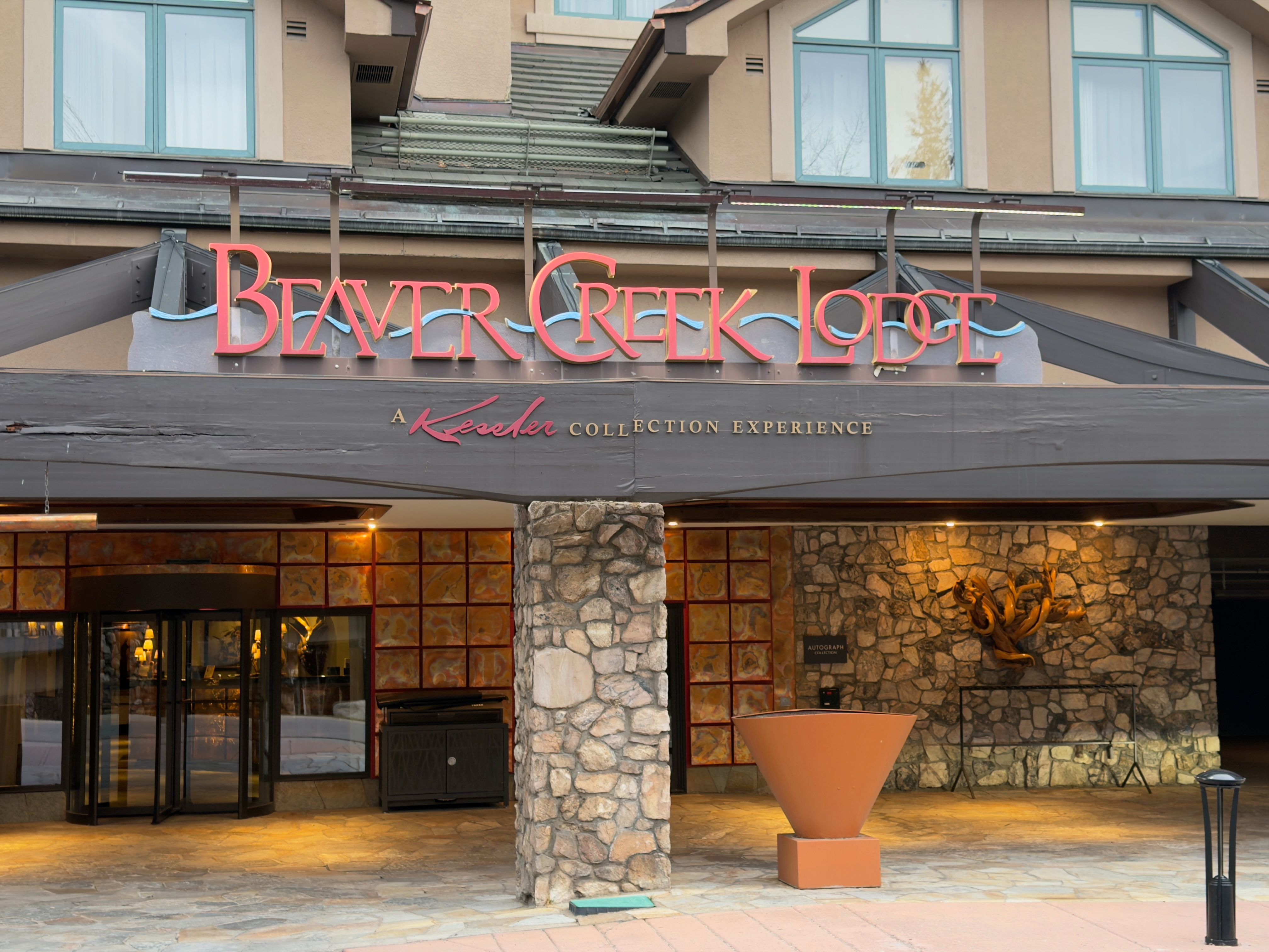 This is an exterior shot of the Beaver Creek Lodge entryway. The entrance features a stone facade, a revolving door, and a prominent sign displaying the lodge's name in red lettering. The overall impression is one of a luxurious and inviting mountain resort.