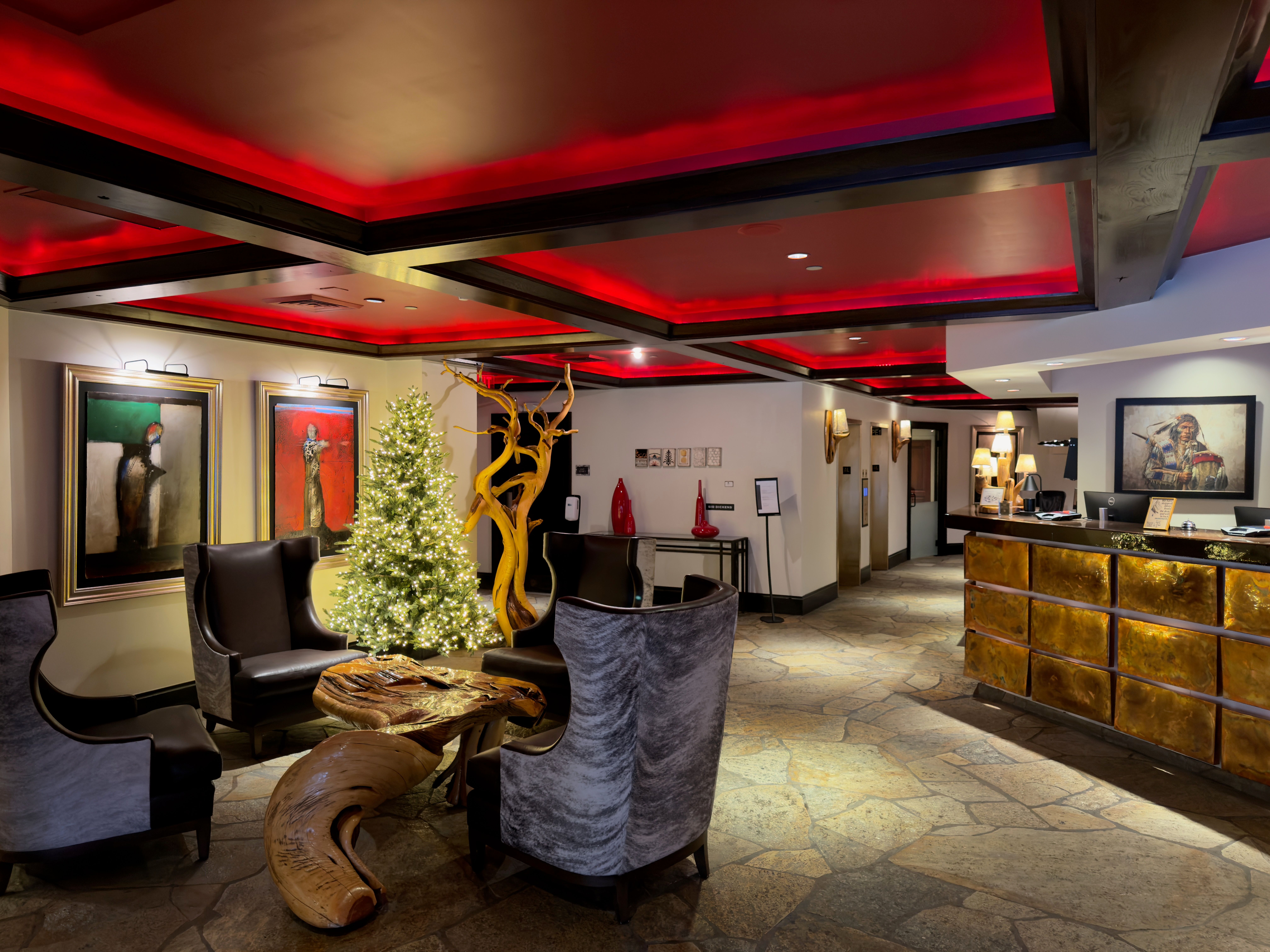 This interior shot showcases a hotel lobby area with a unique blend of rustic and modern design elements. The space features stone flooring, a reception desk with a gold-toned facade, and comfortable seating arrangements around a distinctive wooden table and sculpture. The ceiling is adorned with dark wooden beams and red accent lighting, creating a warm and inviting atmosphere.