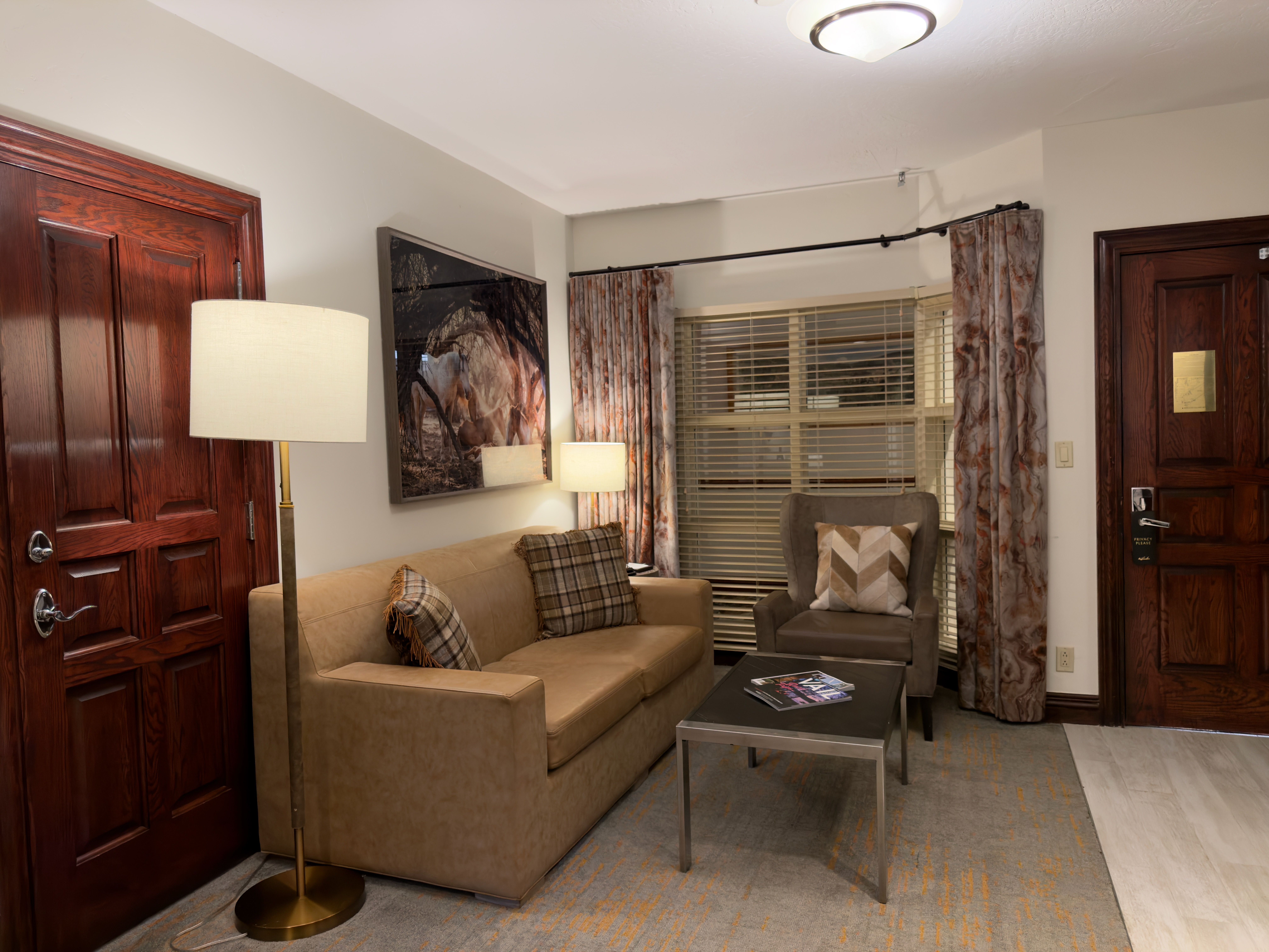 This interior shot showcases a cozy living room with a tan sofa adorned with plaid pillows, complemented by a floor lamp and a side table. A framed artwork hangs above the sofa, and the room features patterned curtains and a comfortable armchair, creating an inviting and stylish space. The room is well-lit, and the overall impression is one of comfort and sophistication.
