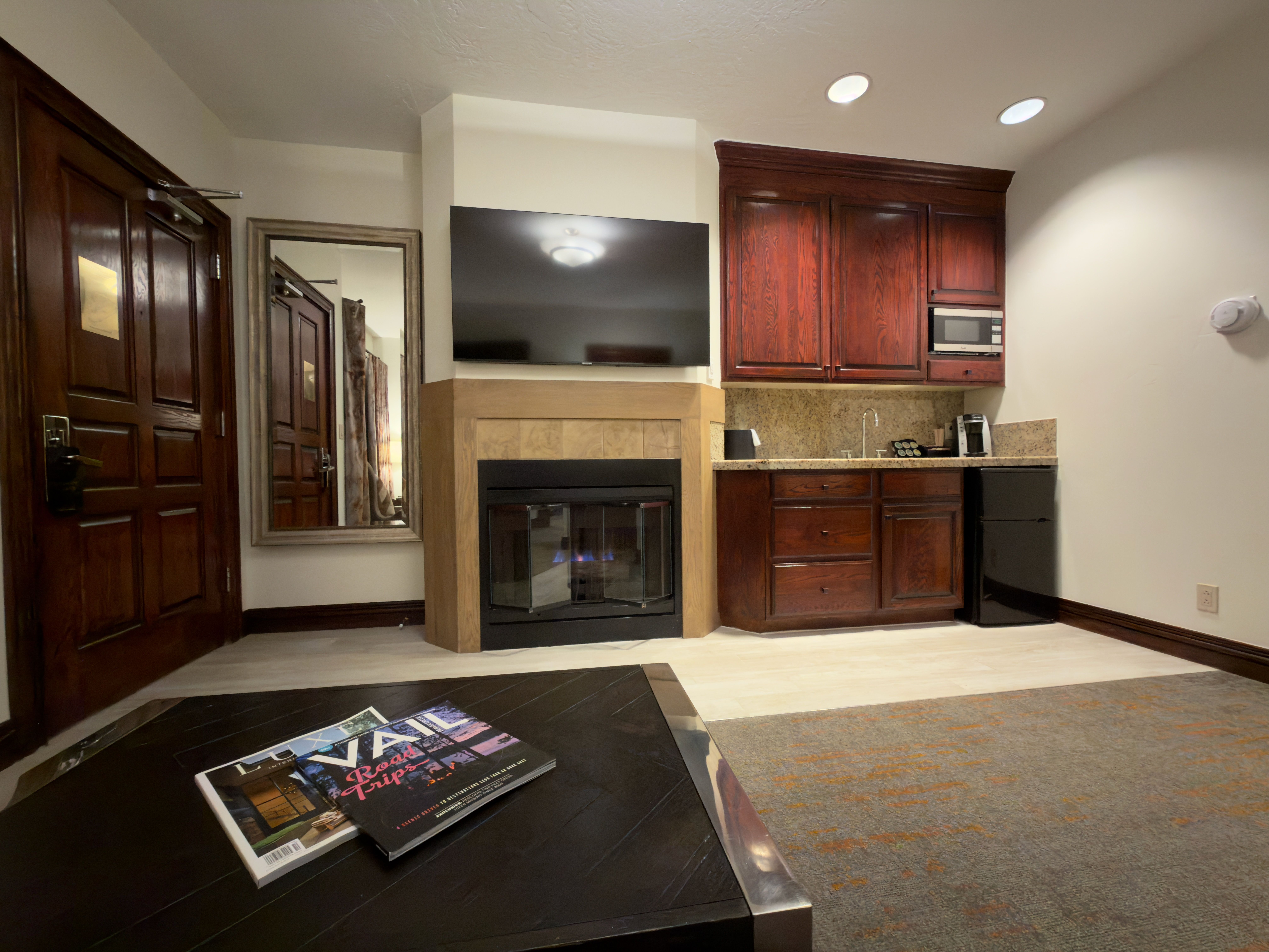 This interior shot showcases a cozy living area with a fireplace and a built-in kitchenette. The room features dark wood accents, including cabinetry and a coffee table, complemented by a neutral color palette. A mirror reflects the room, adding depth, while magazines on the coffee table suggest a relaxed atmosphere.