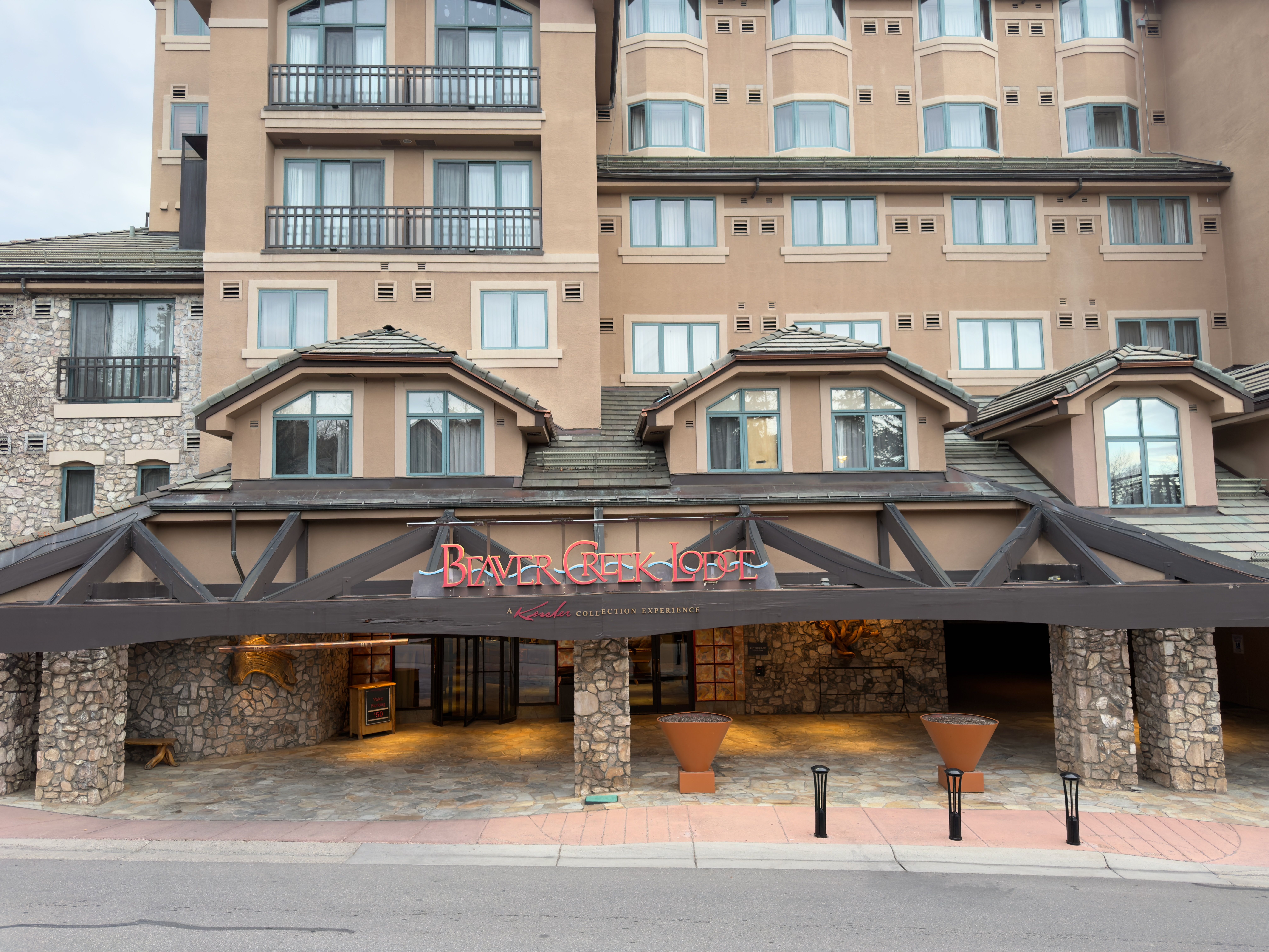 This is an exterior shot of the entryway to Beaver Creek Lodge, showcasing a grand, covered entrance with stone pillars and a dark wood beam structure. The building features a mix of stone and stucco facade, with multiple windows and balconies visible above the entrance. The overall impression is luxurious and inviting, suggesting a high-end resort experience.