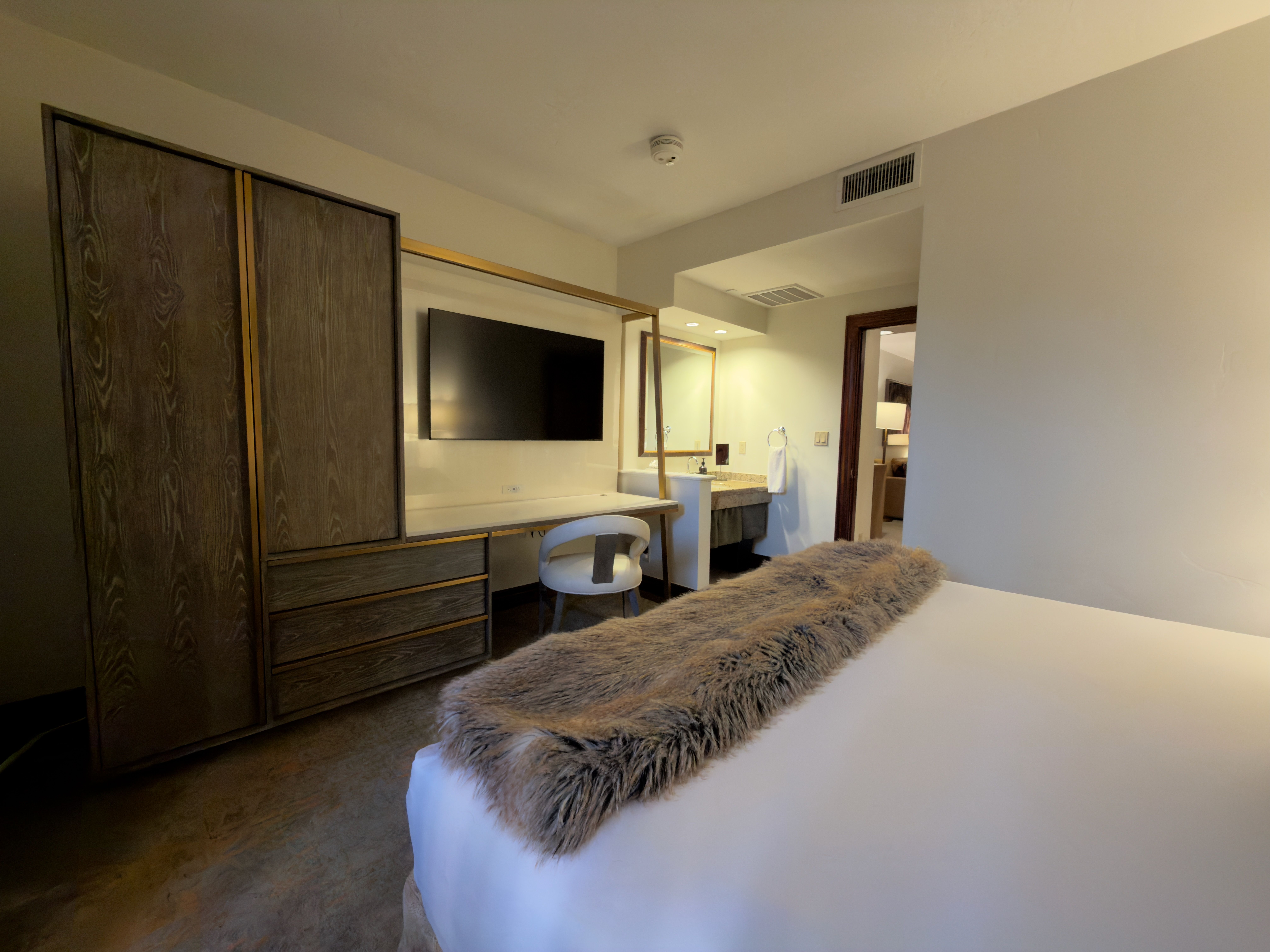 This is a bedroom interior featuring a large bed with a faux fur throw, a built-in wardrobe and desk area with a mounted TV. The room leads into a bathroom, visible through a doorway. The style is modern and comfortable, suitable for a relaxing retreat.