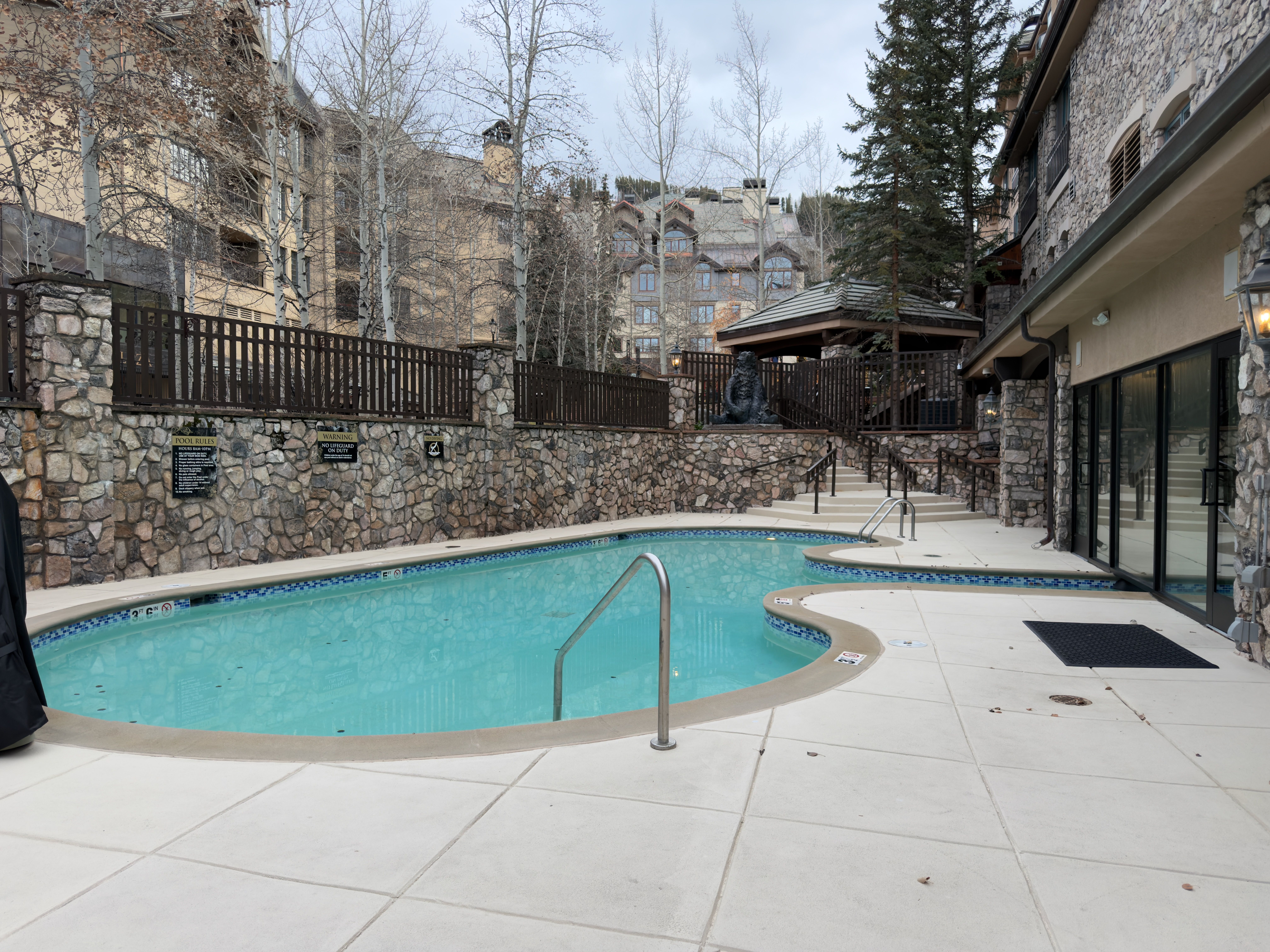 This image showcases an inviting outdoor pool area, featuring a clear blue pool surrounded by a light-colored tiled deck. A stone wall with a wooden fence provides privacy and aesthetic appeal, while a nearby gazebo and building architecture add to the luxurious ambiance. The scene suggests a relaxing and upscale recreational space.