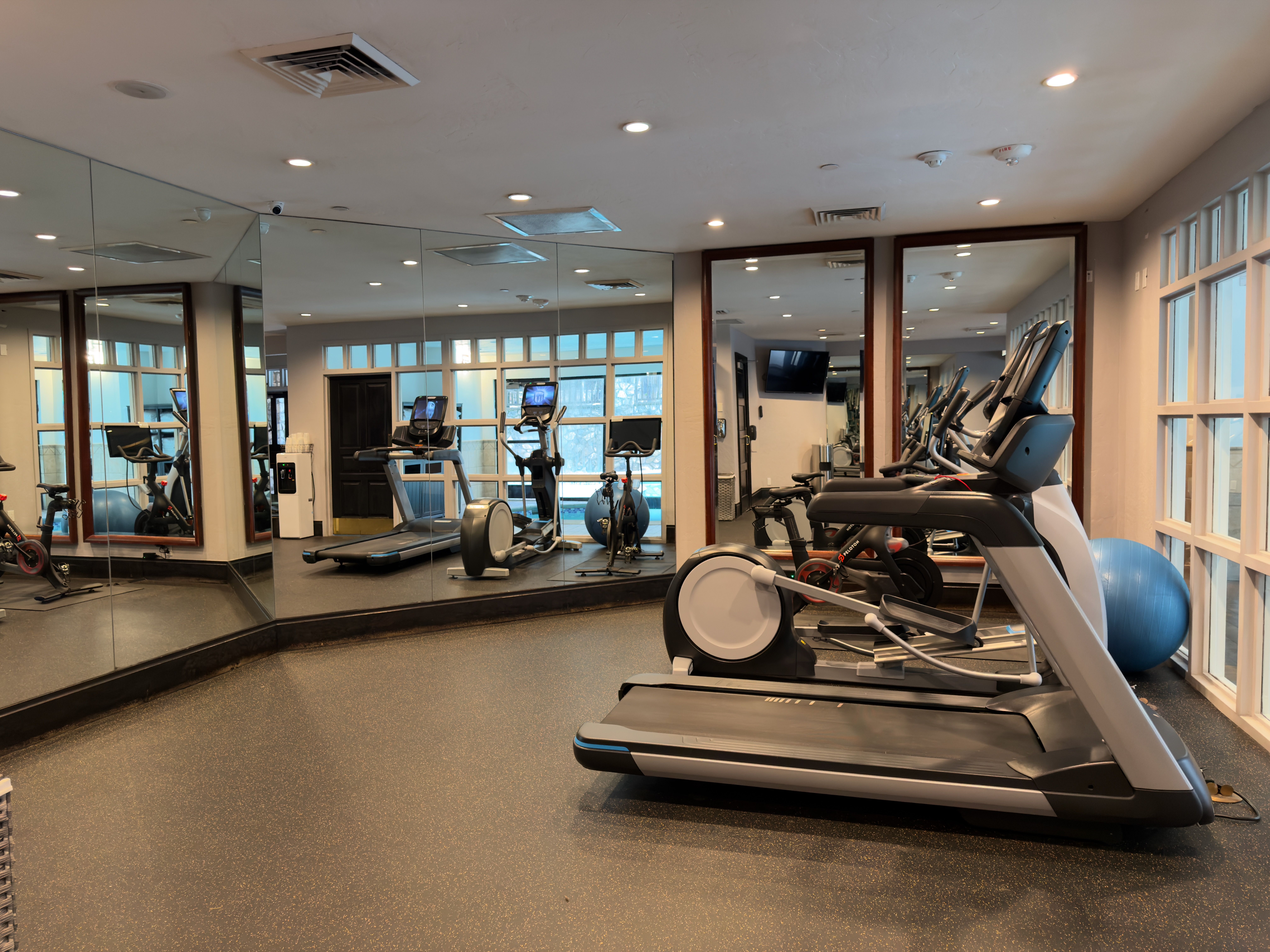 This is a well-equipped home gym featuring treadmills, elliptical machines, and stationary bikes. The room has mirrored walls to enhance the sense of space and allow for form checks during workouts. The flooring is a durable, non-slip material, and the overall impression is clean and functional, perfect for a dedicated fitness space.