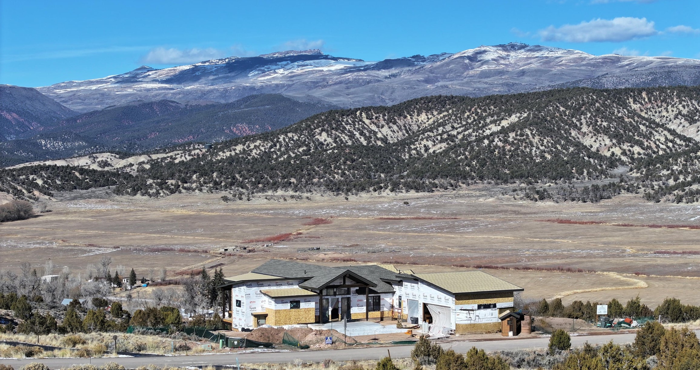 This image captures a wide-angle, elevated perspective of a single-story home currently under construction, set against a dramatic backdrop of expansive, snow-dusted mountains and a vast, open valley. The structure features a modern, multi-gabled roofline and is wrapped in protective white building paper, revealing the underlying wood framing and structural progress. The scene emphasizes the property's remote, scenic location and its integration into the rugged, high-desert landscape.