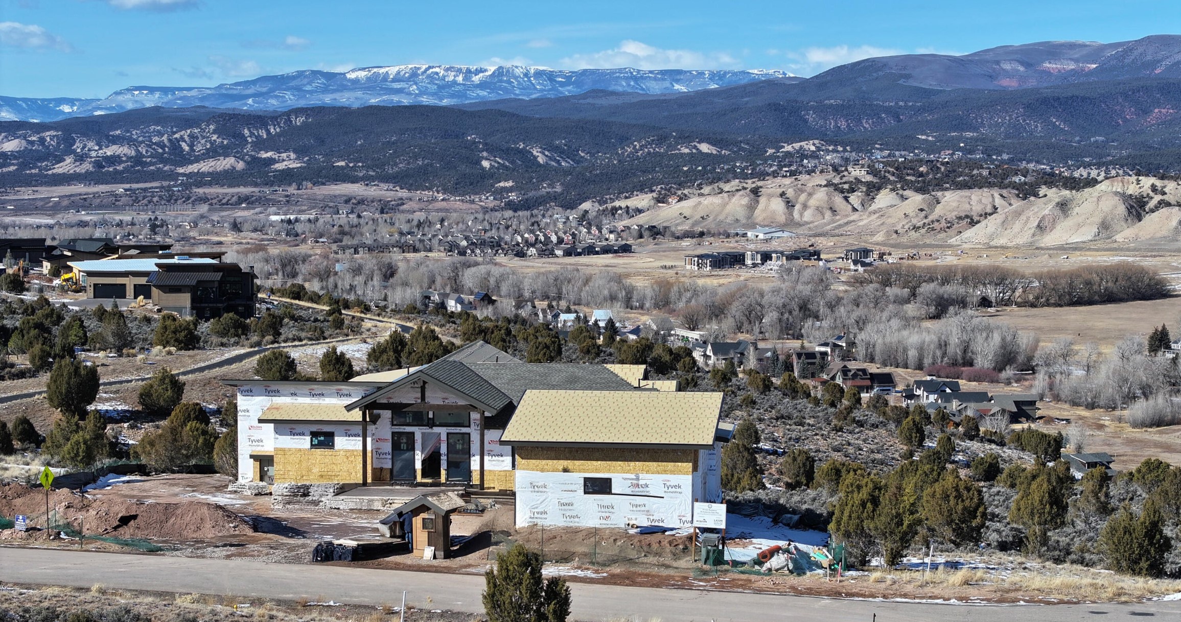 This elevated perspective captures a new construction home in the framing stage, featuring a prominent gabled roofline and Tyvek-wrapped exterior walls. The property is situated in a scenic, mountainous landscape with expansive views of snow-capped peaks and a valley below. The surrounding area includes a mix of established residential homes, sparse vegetation, and open terrain, emphasizing the home's quiet, semi-rural setting.