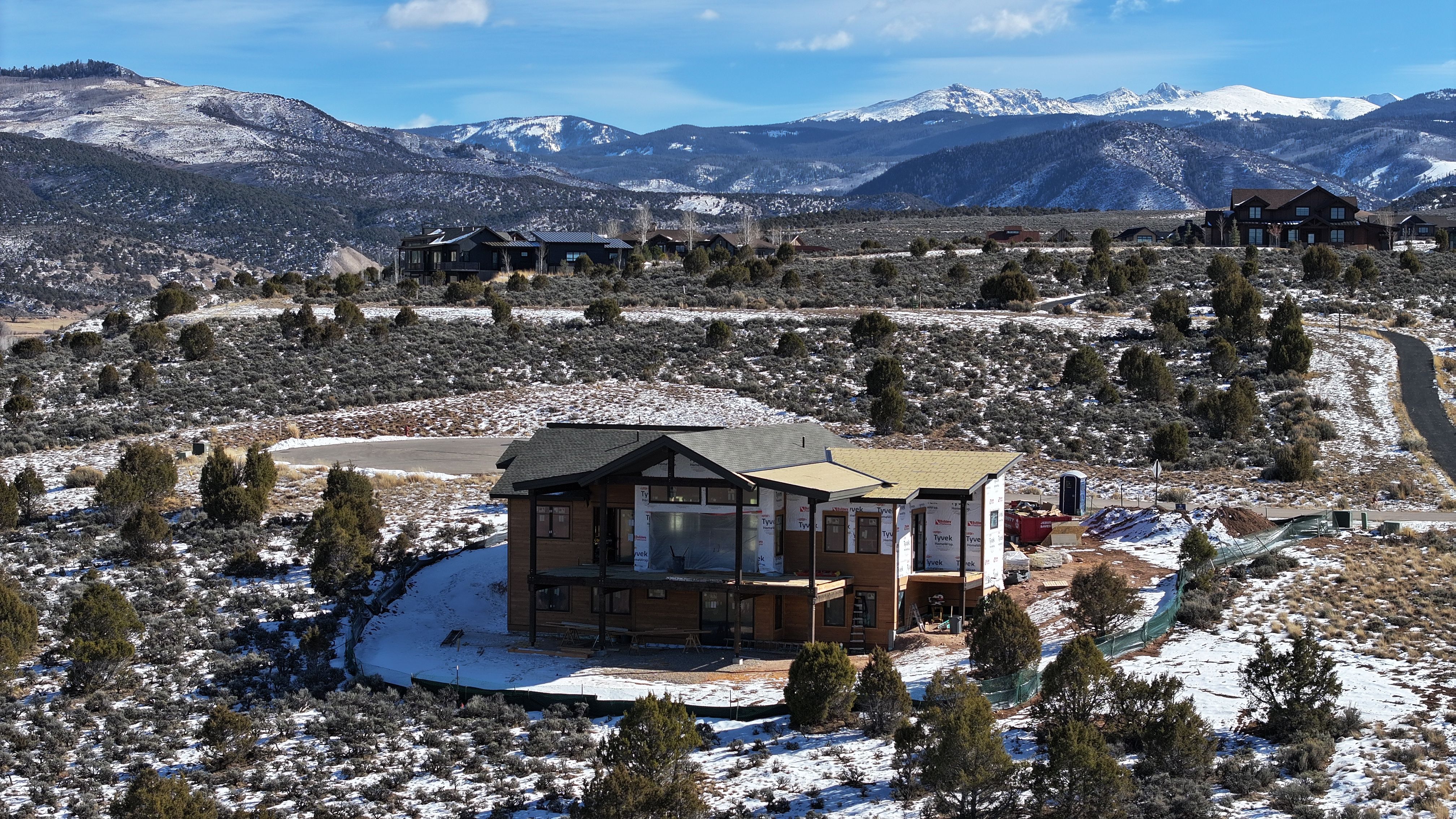 This aerial perspective captures a large, multi-level home under construction, set against a backdrop of snow-dusted mountains and a high-desert landscape. The structure features a prominent gabled roofline, exposed framing, and sections wrapped in protective white house wrap, indicating an ongoing building project. The surrounding terrain is dotted with low-lying shrubs and scattered patches of snow, emphasizing the property's expansive and scenic rural location.