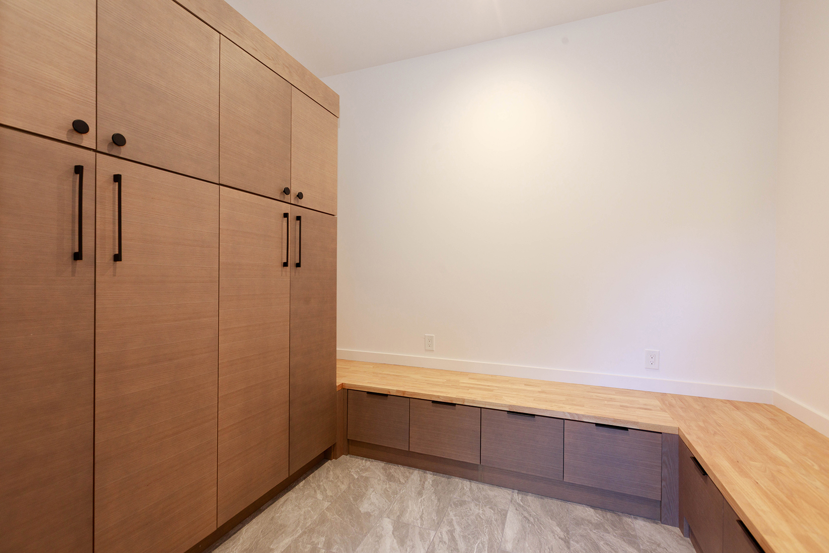 This interior shot showcases a well-organized laundry room featuring floor-to-ceiling wooden cabinets with sleek black hardware. A built-in bench with drawers provides seating and storage, complemented by a light-colored countertop. The room is brightly lit, highlighting the clean lines and modern design.