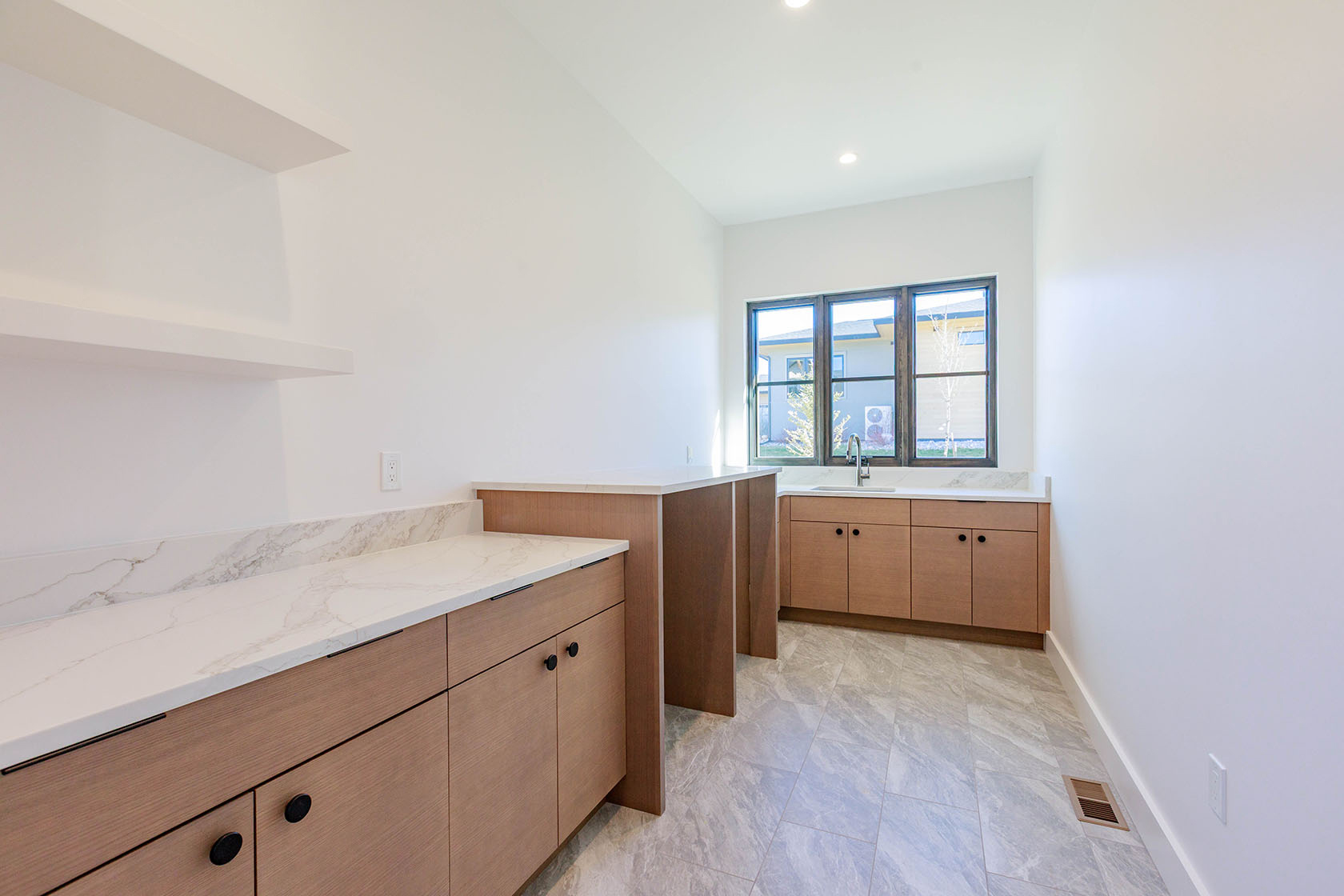 This is an interior shot of a modern pantry. The pantry features light wood cabinetry with black hardware and white marble countertops. A window provides natural light, and the floor is tiled with a gray marble pattern. The overall impression is clean and organized.