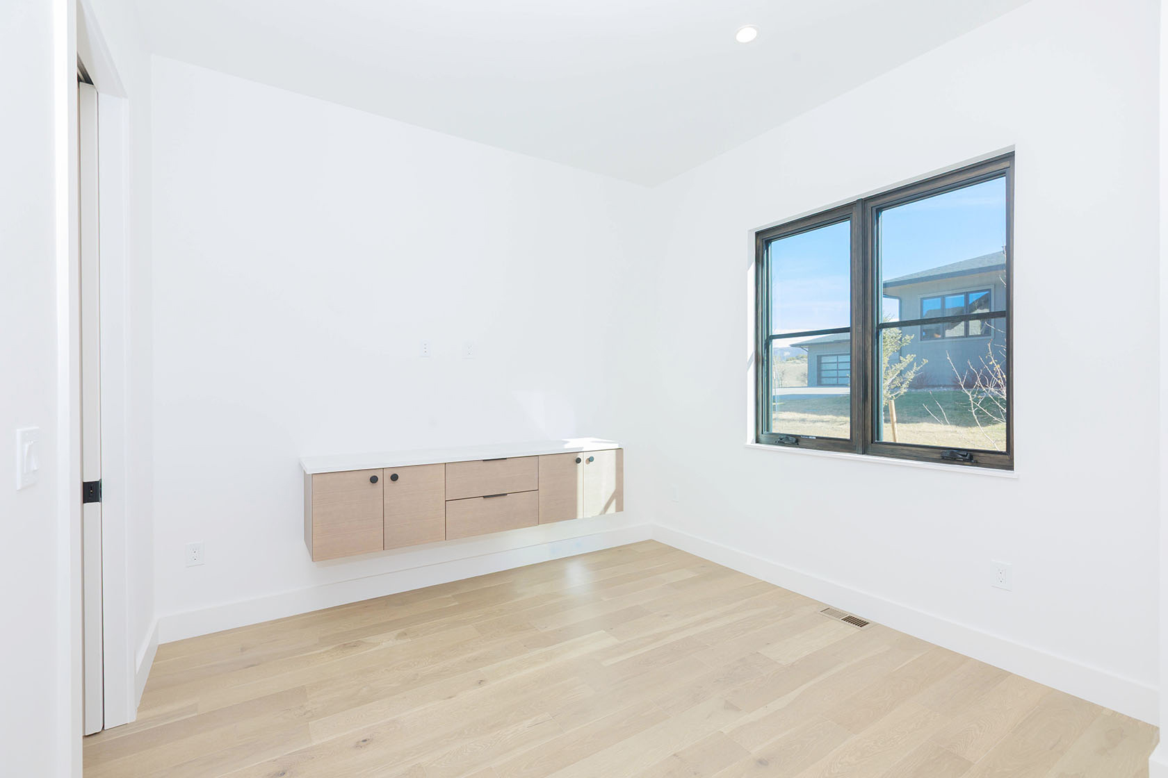 This is a bright, minimalist bedroom featuring light wood flooring and white walls, creating a clean and spacious feel. A floating cabinet with a white countertop is mounted on the wall, and a large window provides natural light and a view of the outside. The room is simple yet elegant, perfect for a guest bedroom or office space.