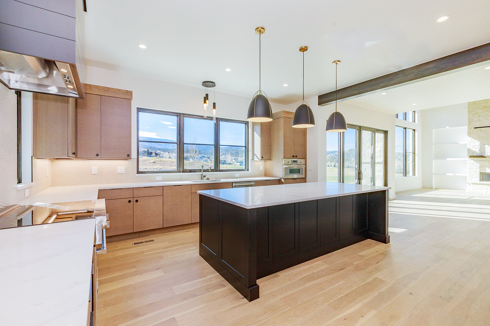 This is a bright and modern kitchen featuring light wood cabinetry, a dark-colored island with a white countertop, and stainless steel appliances. The kitchen is well-lit with pendant lighting and recessed lighting, and large windows offer a view of the landscape. The open floor plan seamlessly connects the kitchen to the living area, creating a spacious and inviting atmosphere.