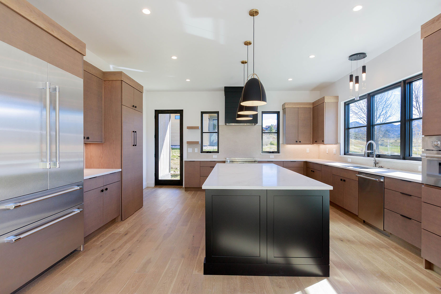 This is a bright and modern kitchen featuring light wood flooring, two-tone cabinetry with a dark island, and stainless steel appliances. The kitchen is well-lit with recessed lighting and pendant lights over the island, and natural light streams in through a large window. The open layout and clean design create a welcoming and functional space.