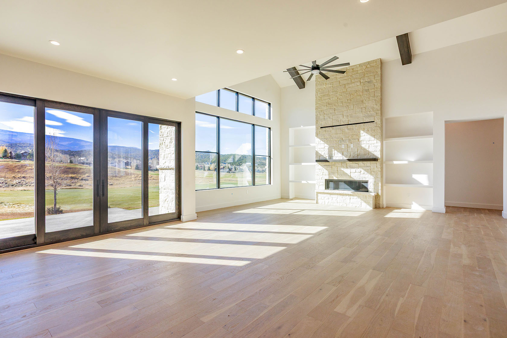 This is a spacious living room featuring a modern design with a stone fireplace and built-in shelving. Large windows and sliding glass doors provide ample natural light and views of the surrounding landscape. The light wood flooring and neutral color palette create a bright and airy atmosphere.
