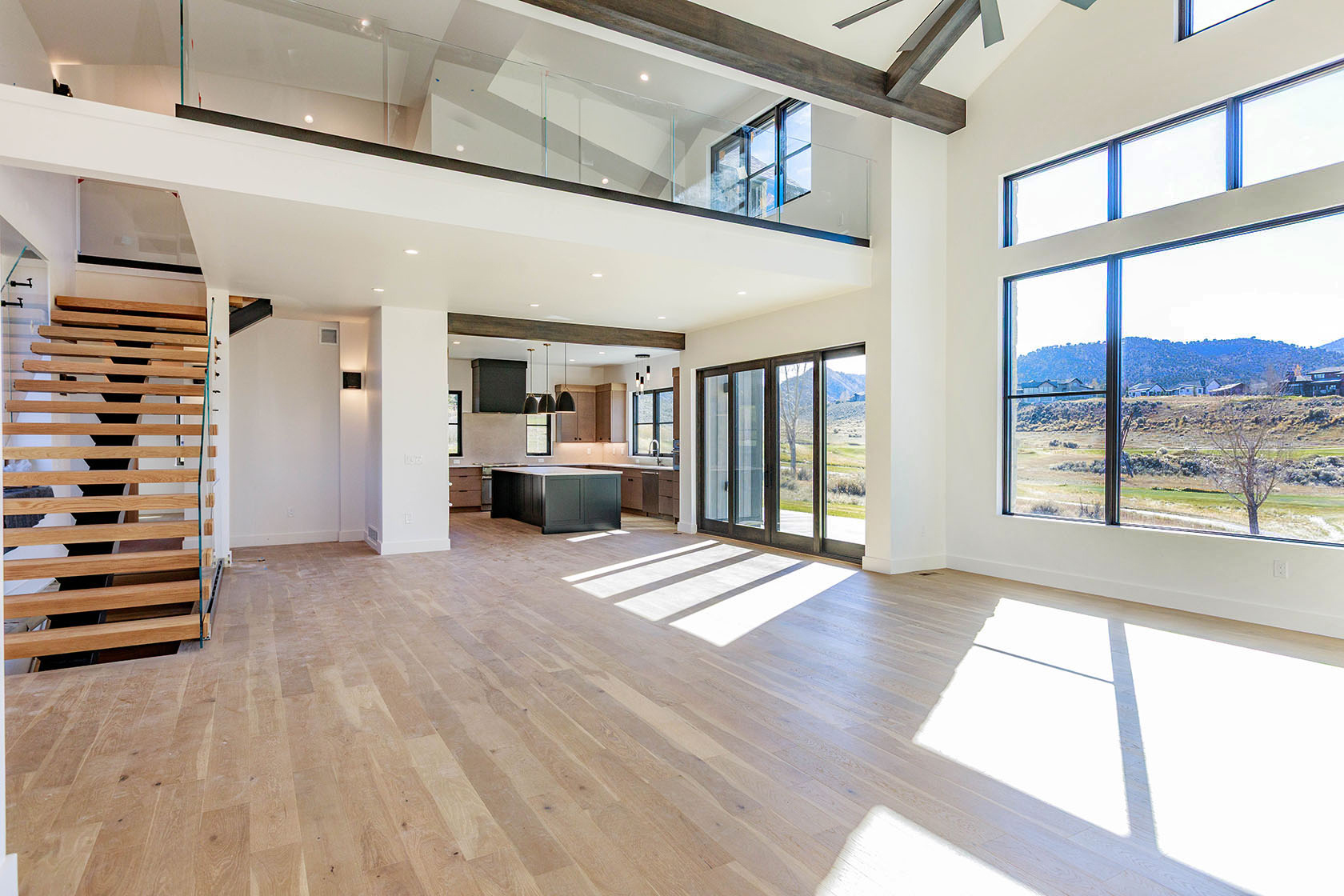 This is a spacious, modern living room with an open floor plan that seamlessly integrates with the kitchen. The room features large windows offering ample natural light and views of the landscape. A striking staircase with open treads and glass railings adds a contemporary touch, while wooden beams provide a rustic contrast. The light wood flooring and neutral color palette create a bright and airy atmosphere.
