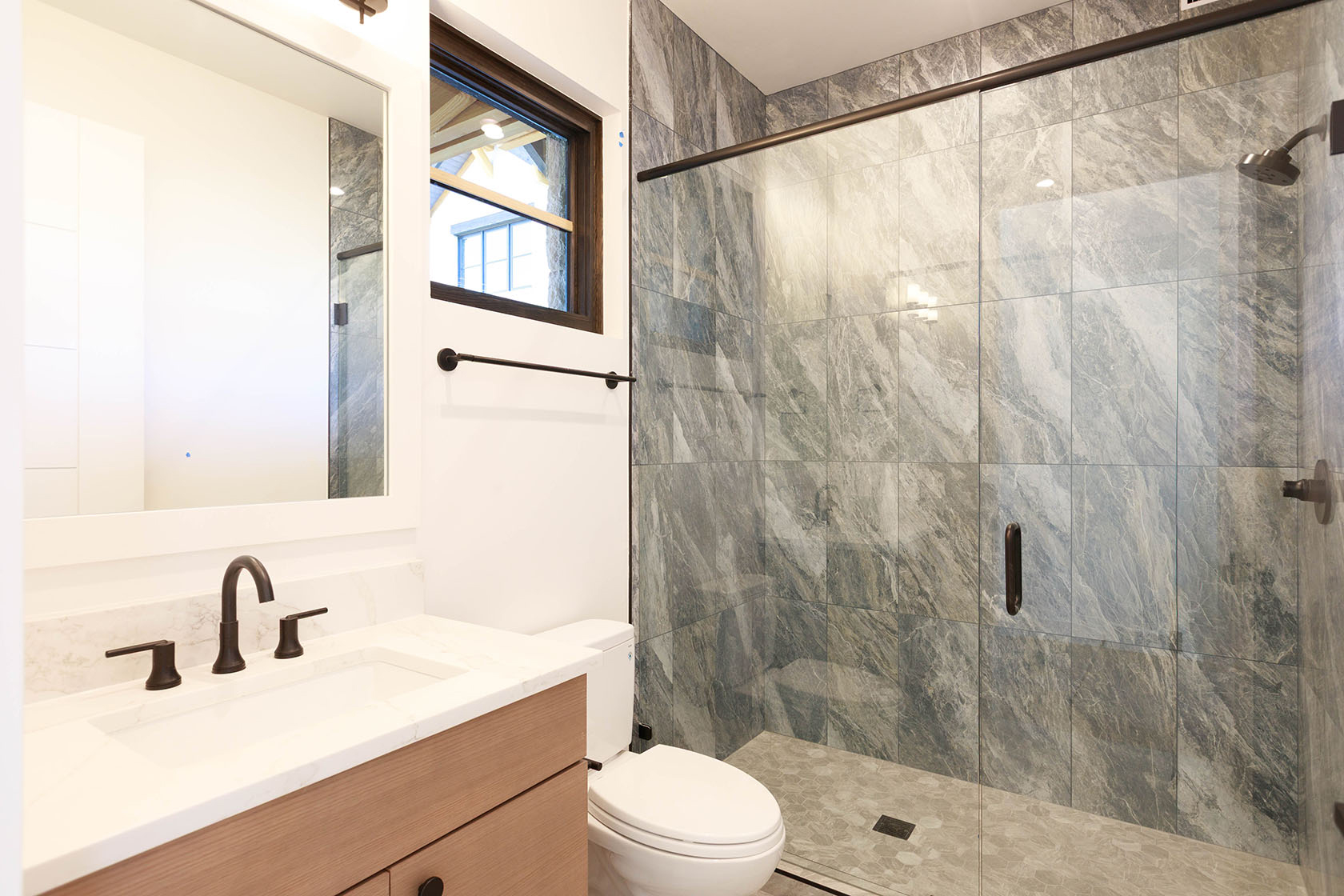 This is a well-lit bathroom featuring a modern design. The vanity has a light wood cabinet with a white countertop and dark fixtures. A glass-enclosed shower with gray marble-like tiles adds a luxurious touch, and a toilet is positioned next to the vanity.