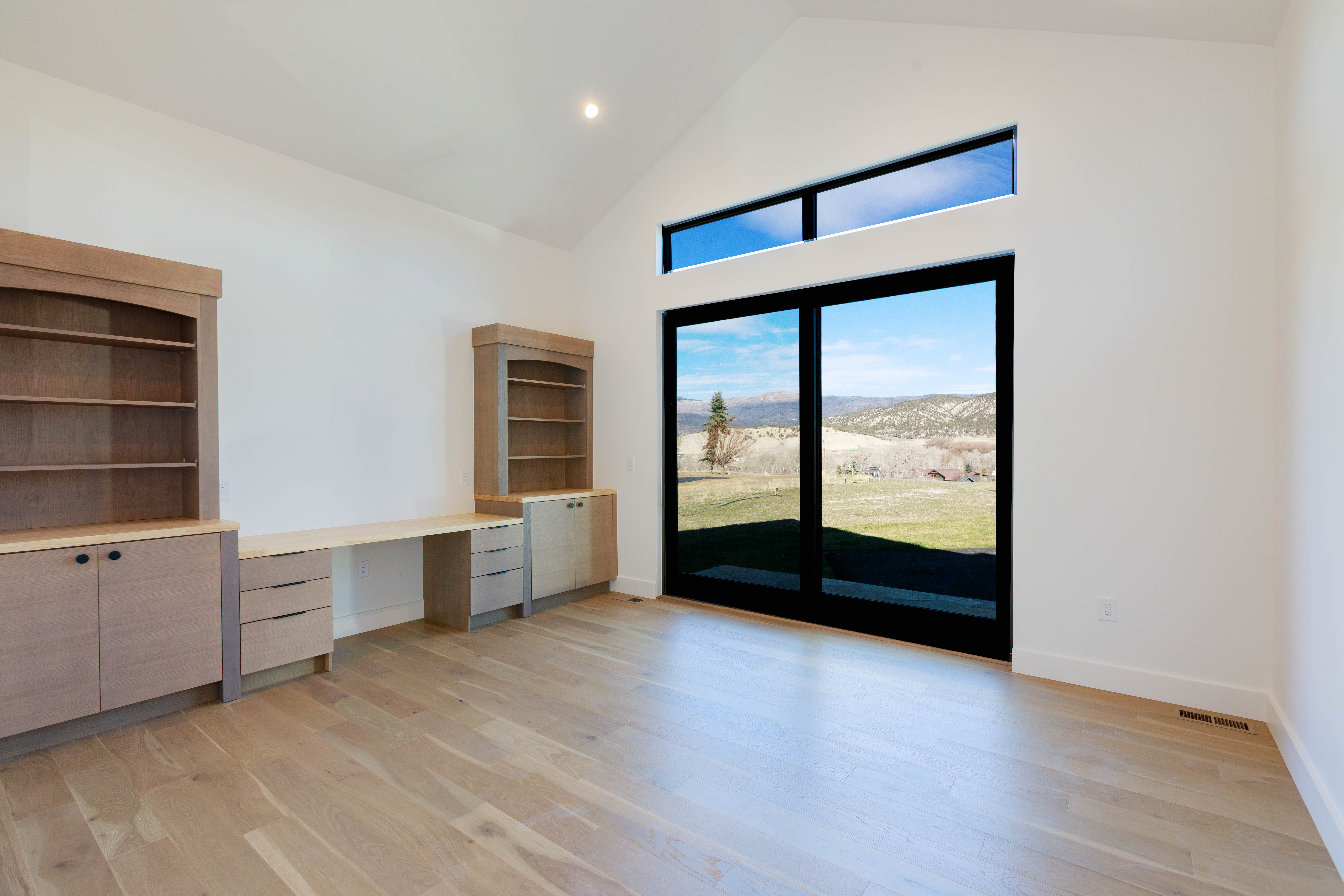 This is a bright and spacious office or study featuring custom built-in cabinetry and a desk area. The room has light hardwood flooring and white walls, creating a clean and modern aesthetic. A large sliding glass door provides ample natural light and a view of the outdoor landscape, enhancing the room's appeal.